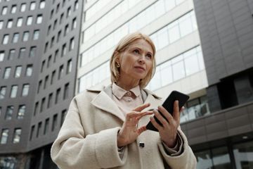 Woman in beige coat using a smartphone in front of a modern building.