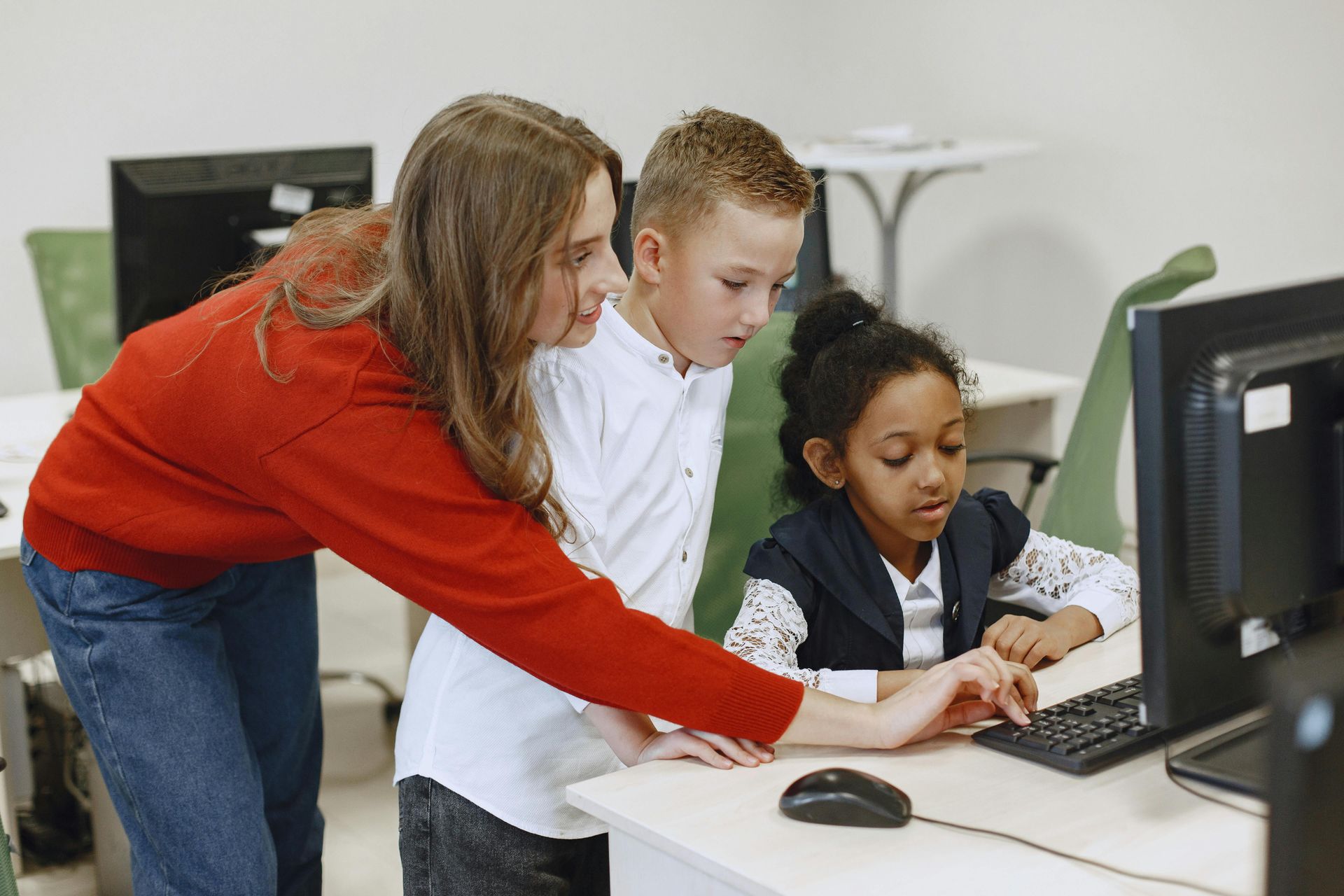 Teacher points at a computer screen with two students in a classroom, light-colored room.
