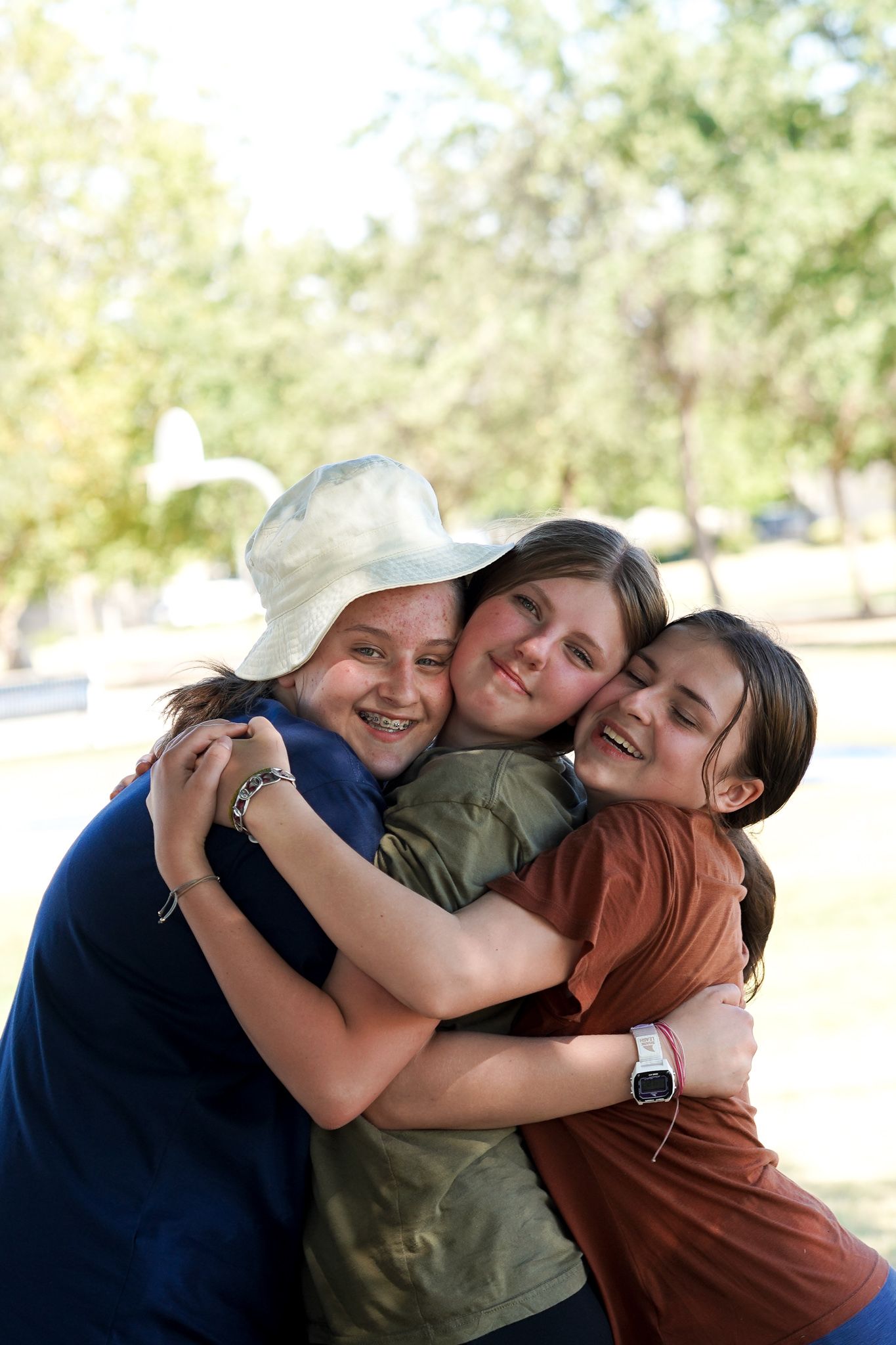 Three young girls are hugging each other in a park.