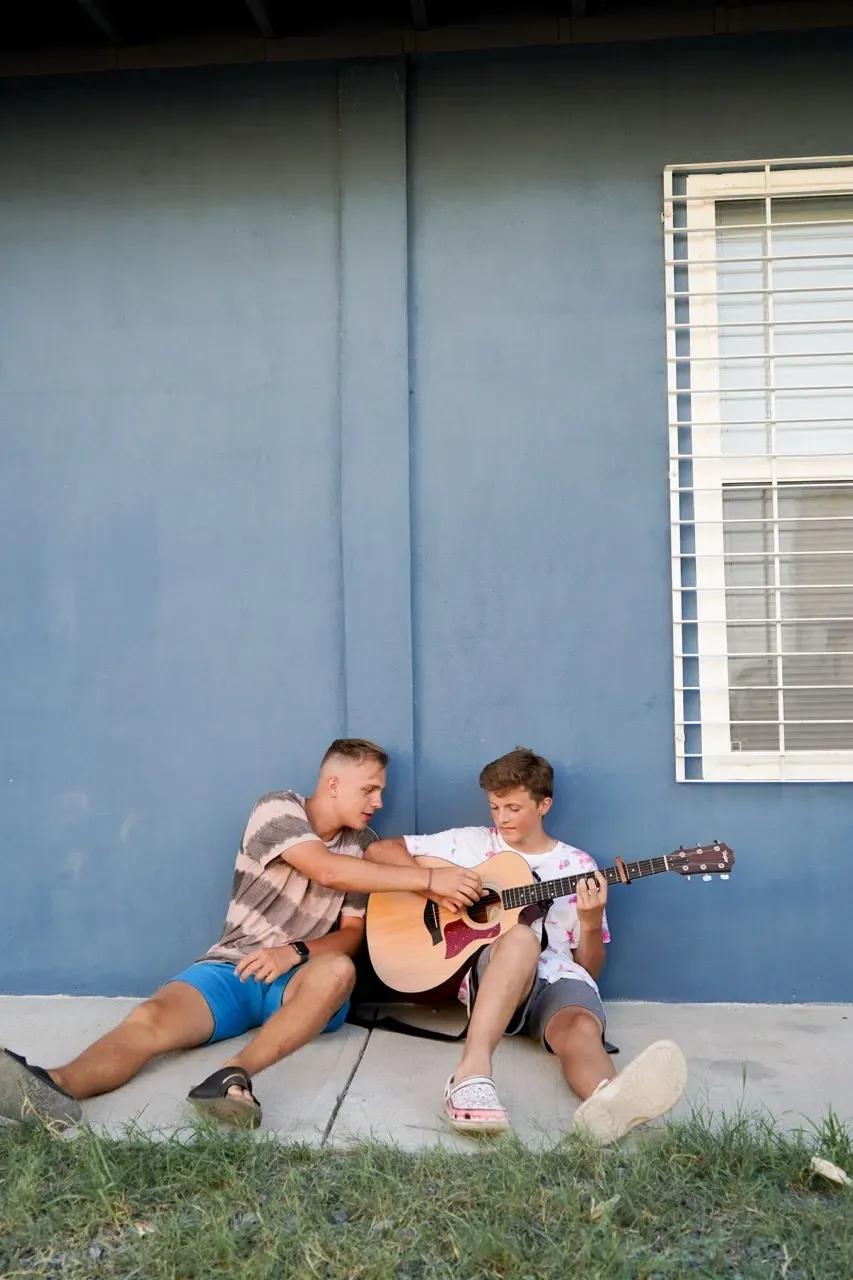 Two men are sitting on the sidewalk playing guitars.