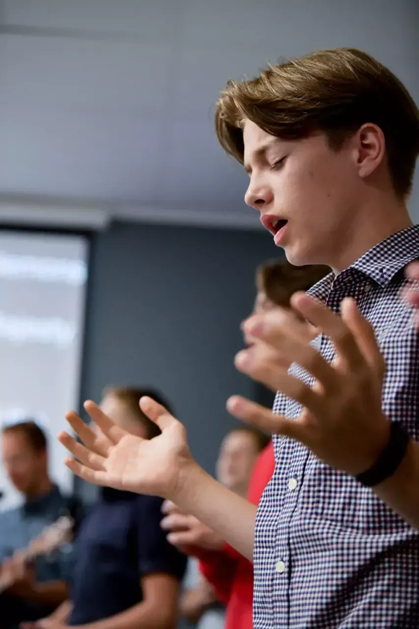 A boy in a plaid shirt is standing in front of a group of people with his hands outstretched.