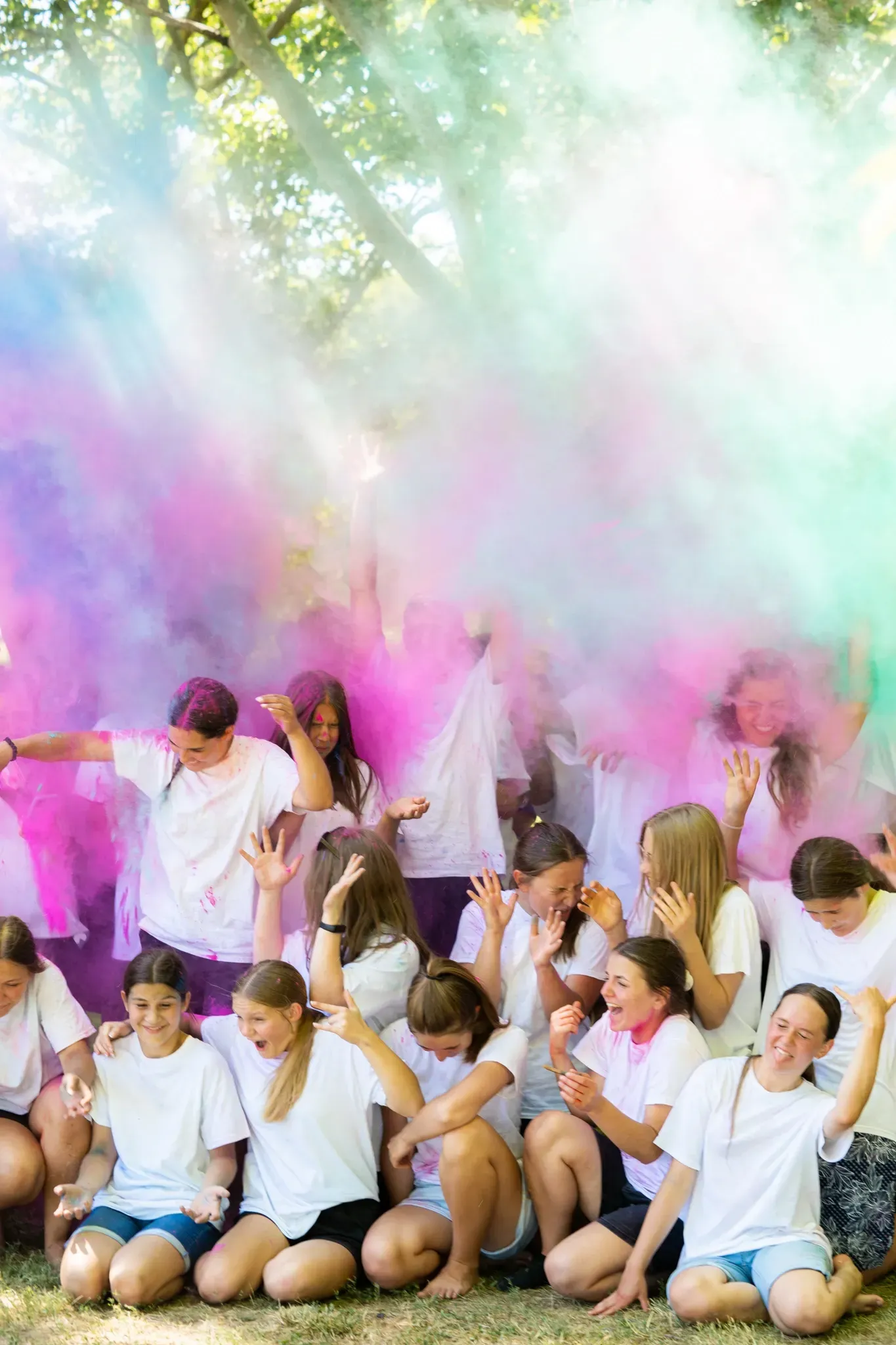 A group of young people are sitting in the grass surrounded by colored smoke.