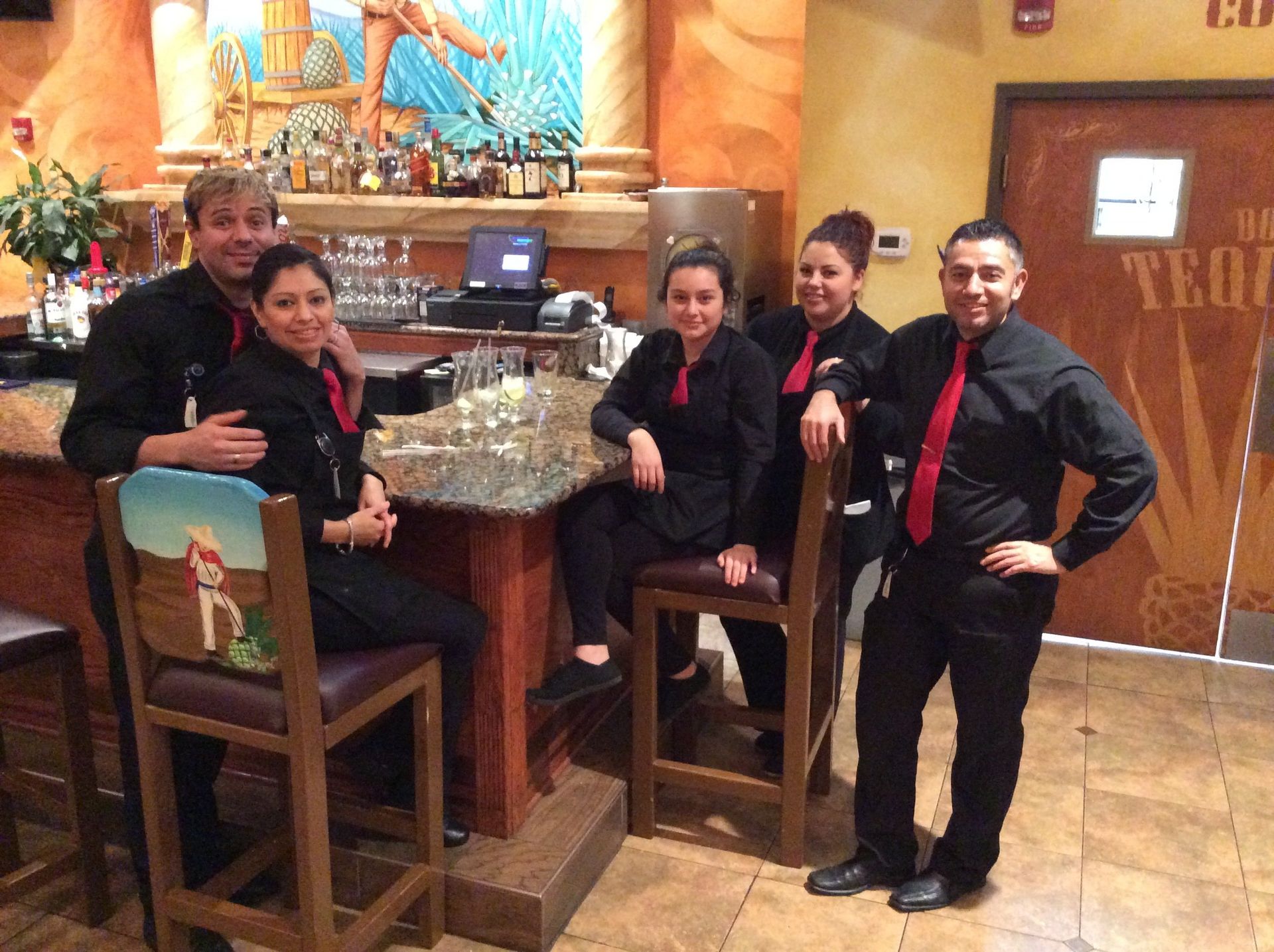 Restaurant staff posing at a bar. They wear black shirts and red ties. The bar has a decorative painting.