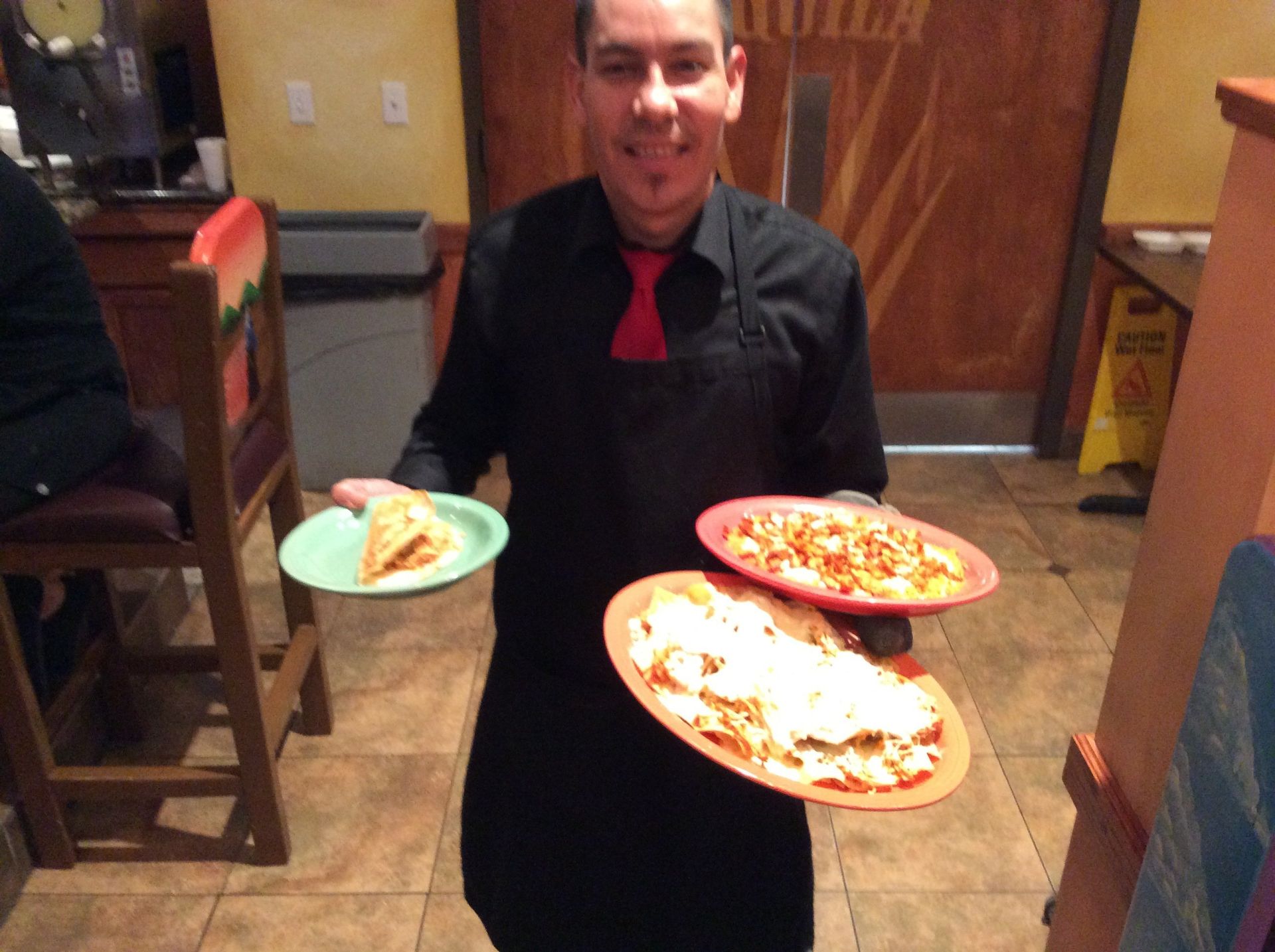 Server carrying three plates of food in a restaurant. He wears a black uniform and red tie, smiling.