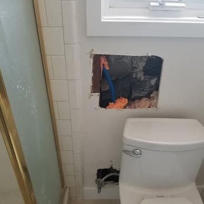 Bathroom wall cut open above toilet, exposing pipes and insulation near a frosted shower door.