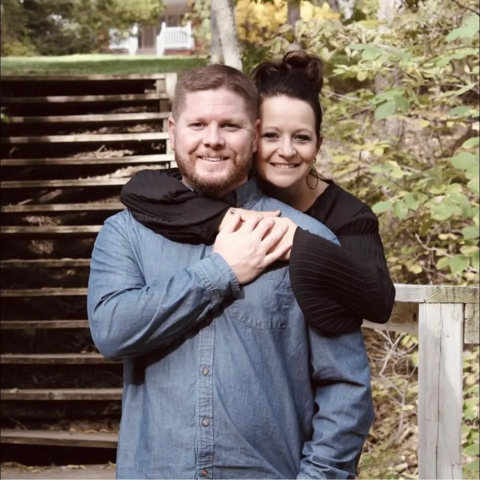 Couple smiling and embracing outdoors near wooden stairs and foliage.