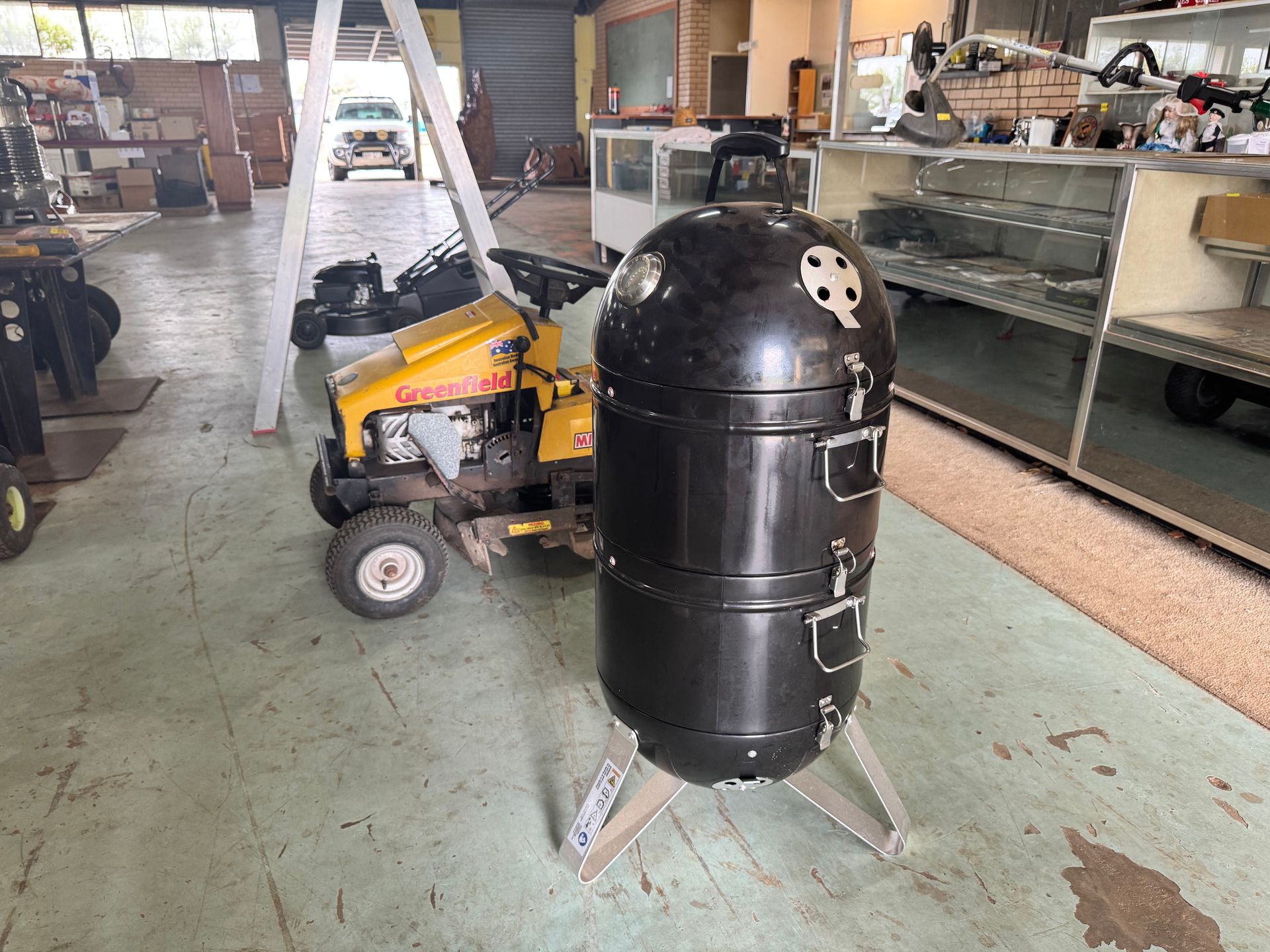 Black Smoker Grill Next to A Yellow Riding Lawnmower Inside a Storage Building — Bundaberg Auction Centre in Gooburrum, QLD