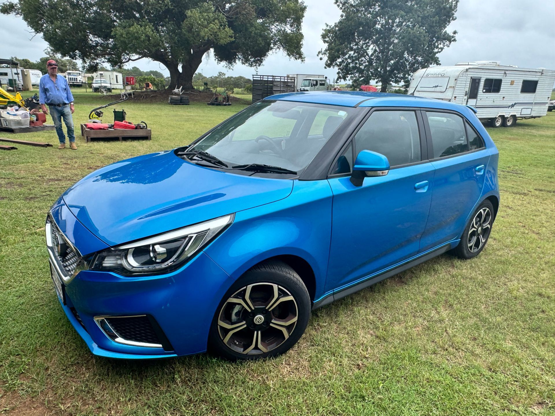 Blue Mg3 Hatchback Parked on Grass; Man in Background Near Equipment and R Vs — Bundaberg Auction Centre in Gooburrum, QLD
