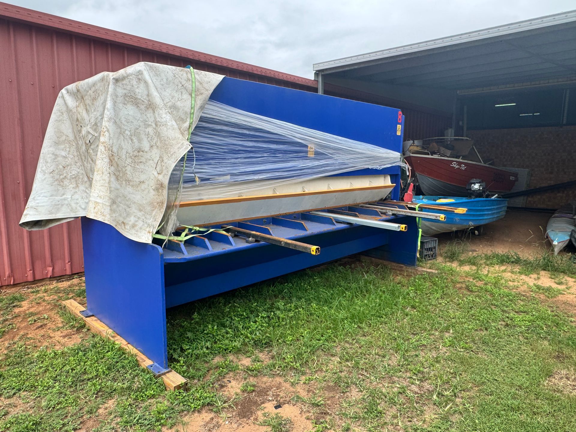 Blue Metal Shear Machine Under a Tarp on Grass, Boats in The Background — Bundaberg Auction Centre in Gooburrum, QLD