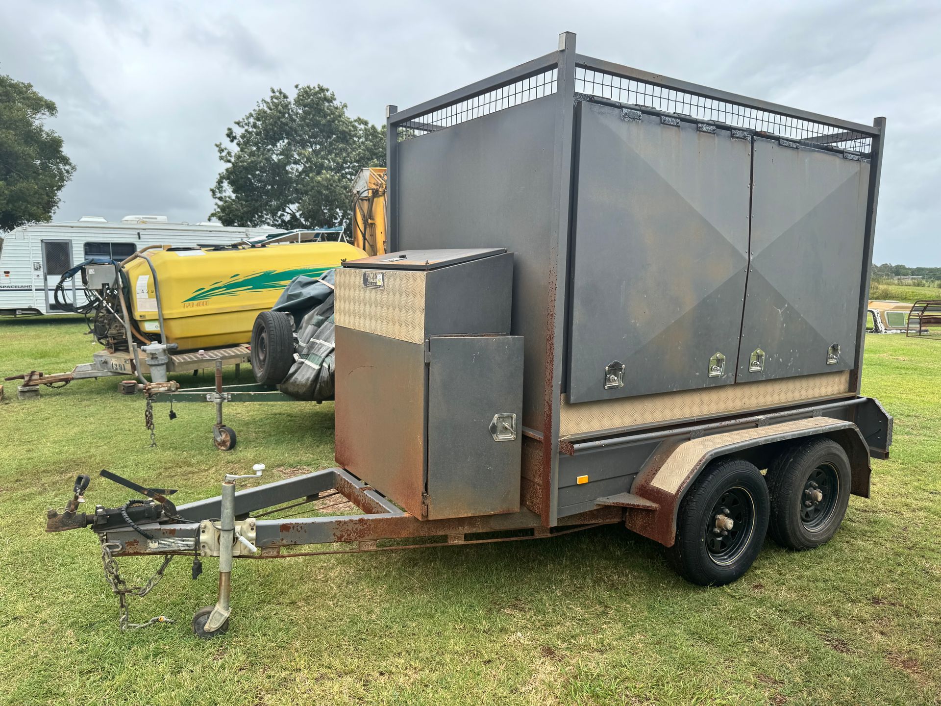 A Weathered Gray Trailer with Cage, Storage, and Dual Wheels, Parked on Grass — Bundaberg Auction Centre in Gooburrum, QLD