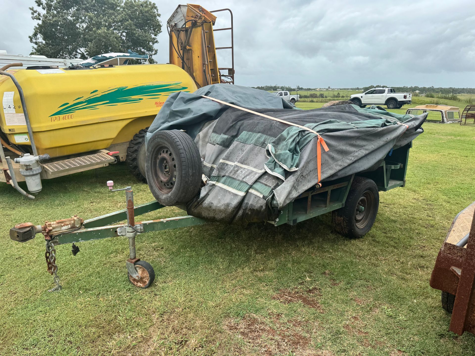 Green Utility Trailer with Spare Tire and Tarp, Parked on Grass.  a Yellow Tank Truck Is Next to It — Bundaberg Auction Centre in Gooburrum, QLD