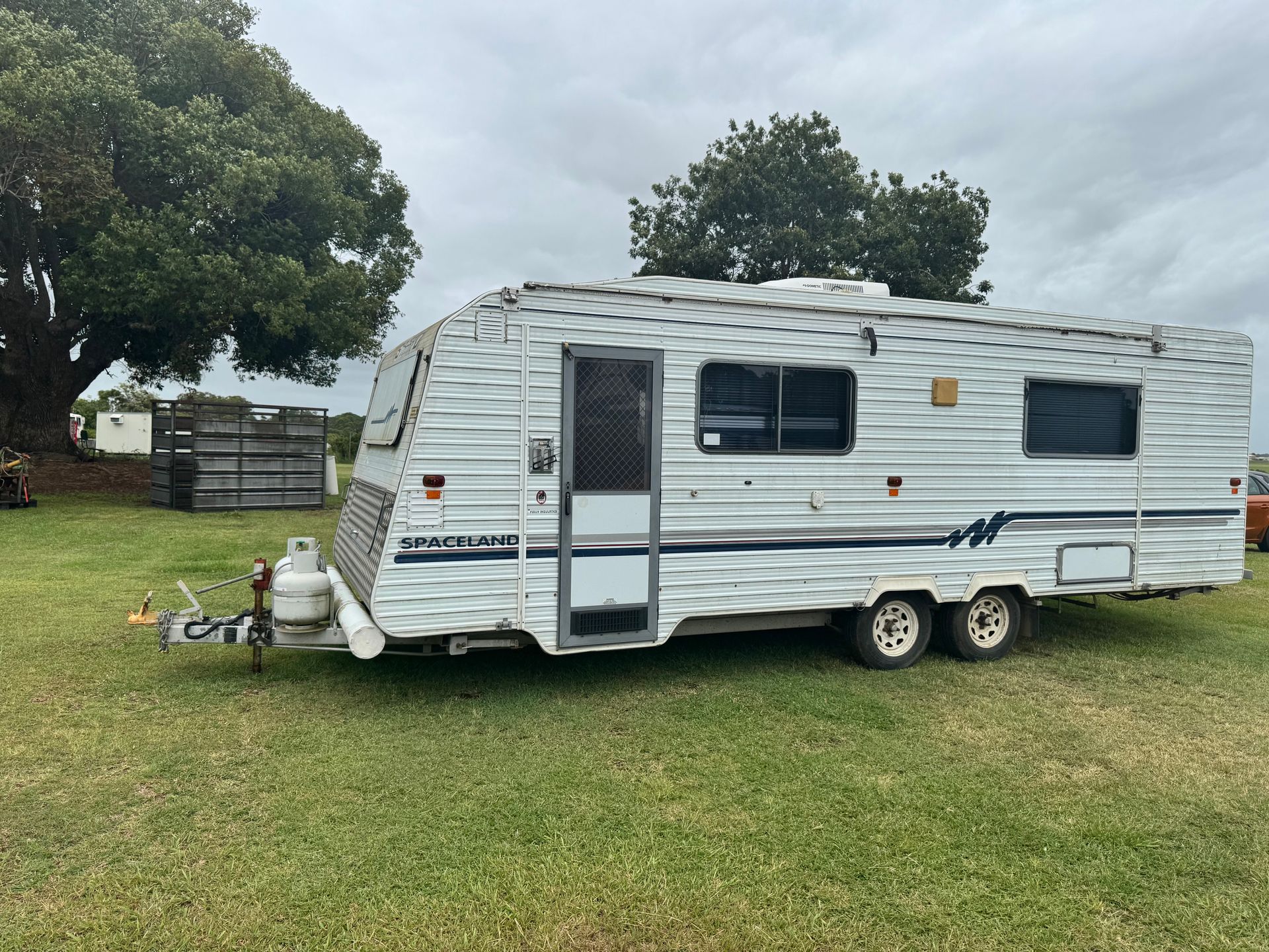 White and Blue Caravan Parked on Grass with A Gas Tank and Trailer Hitch Visible — Bundaberg Auction Centre in Gooburrum, QLD