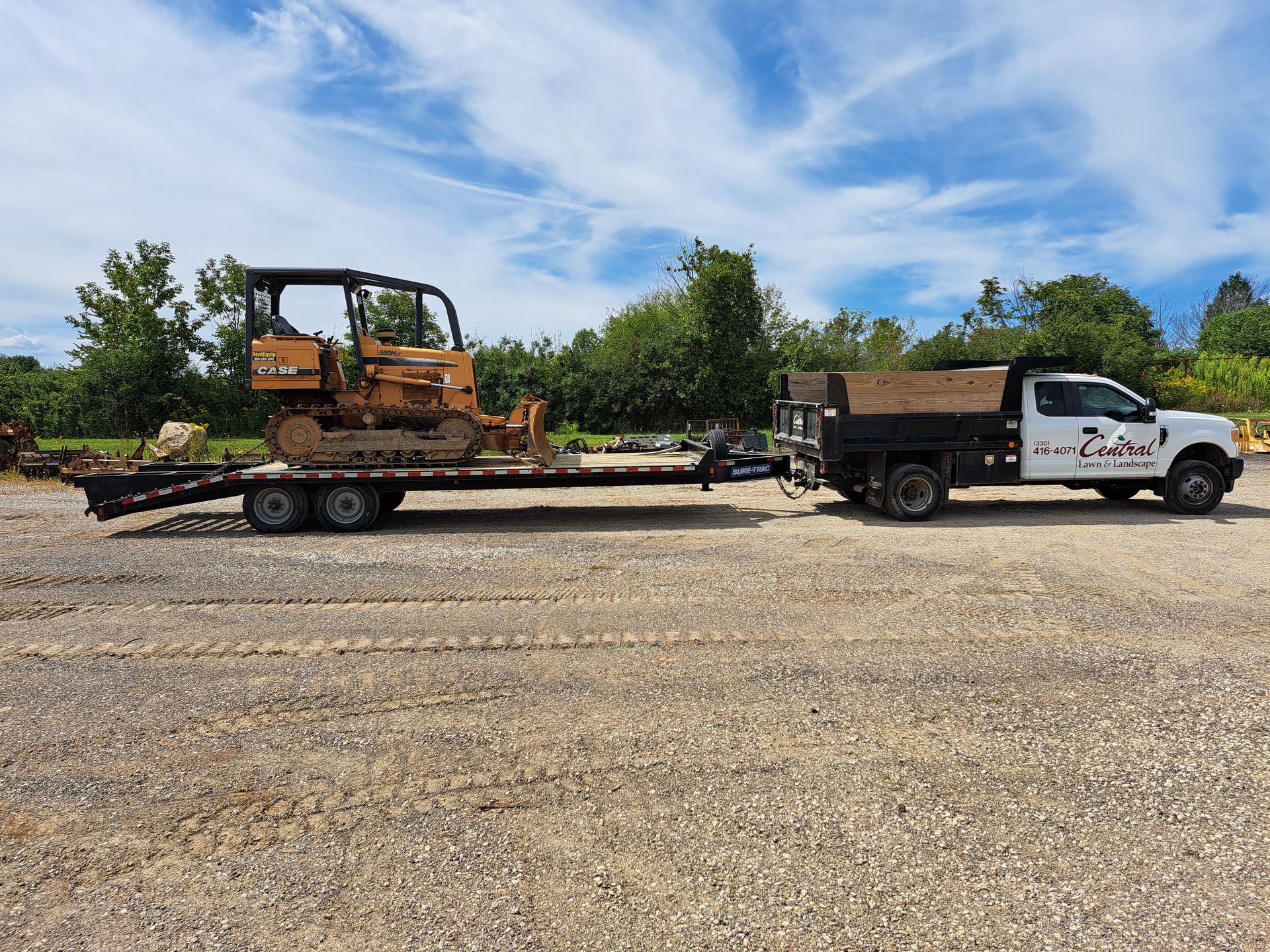 Truck towing a trailer with a bulldozer on it, parked on gravel, under a blue sky.