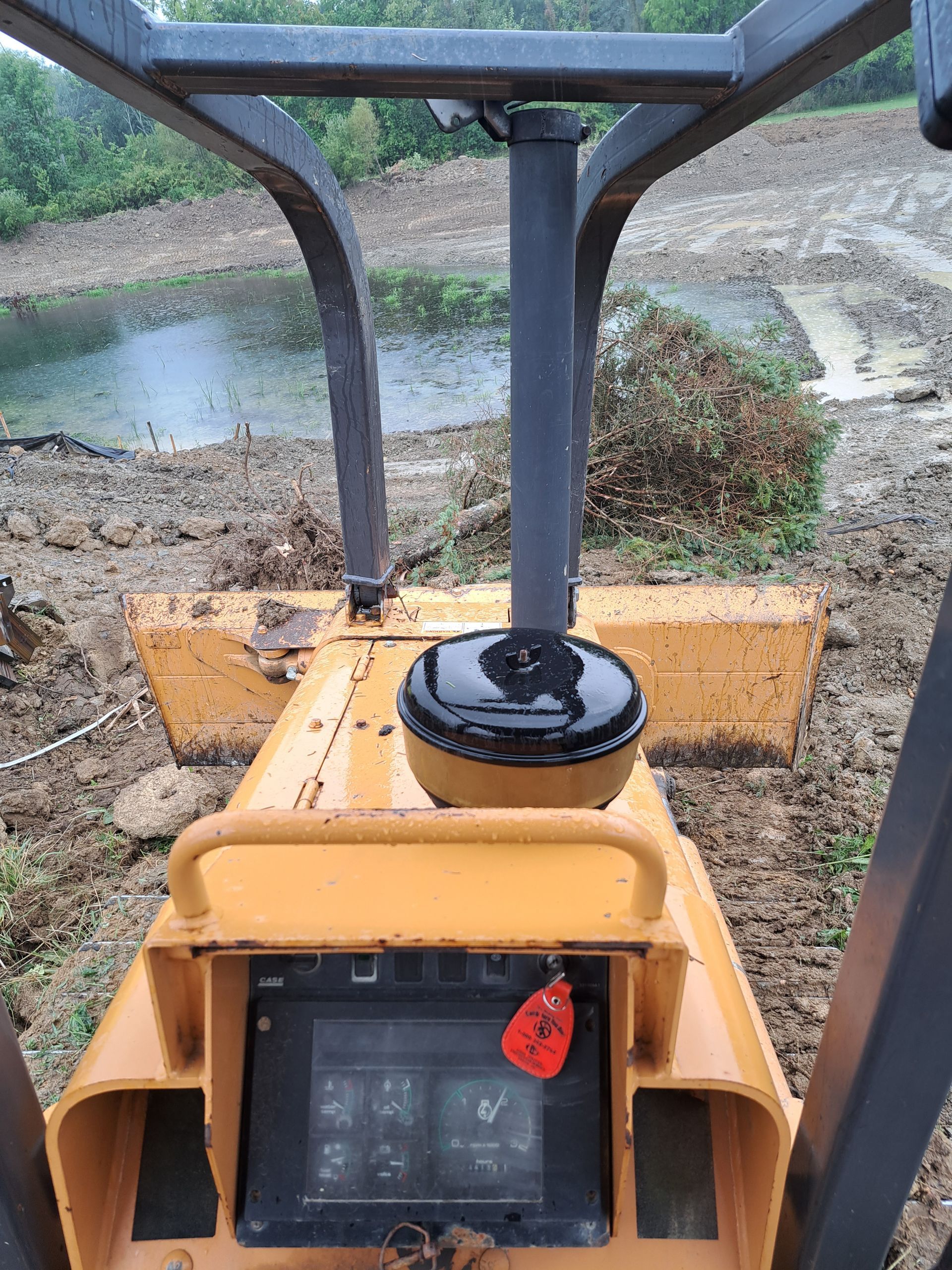 View from inside a yellow bulldozer cab, looking at the blade and a muddy worksite with water in the background.
