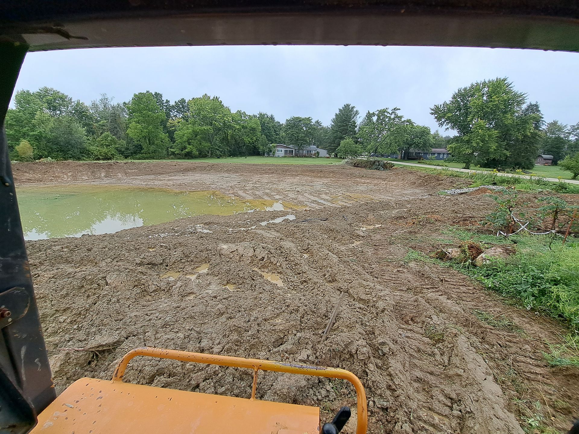 View from inside a bulldozer cab, looking at a muddy construction site near trees and a pond.