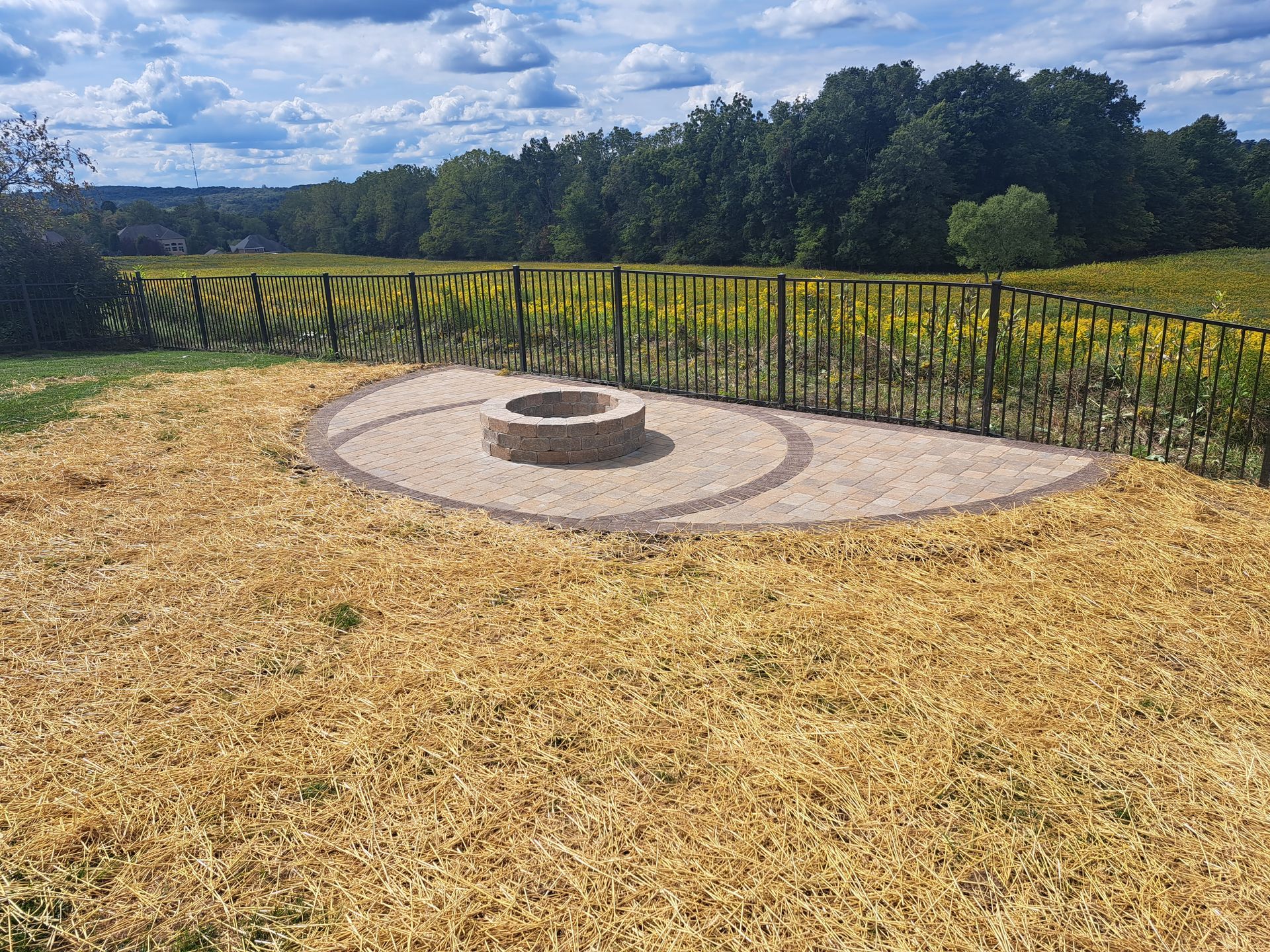 Fire pit area with gravel, stone, and a black metal fence, overlooking a field.
