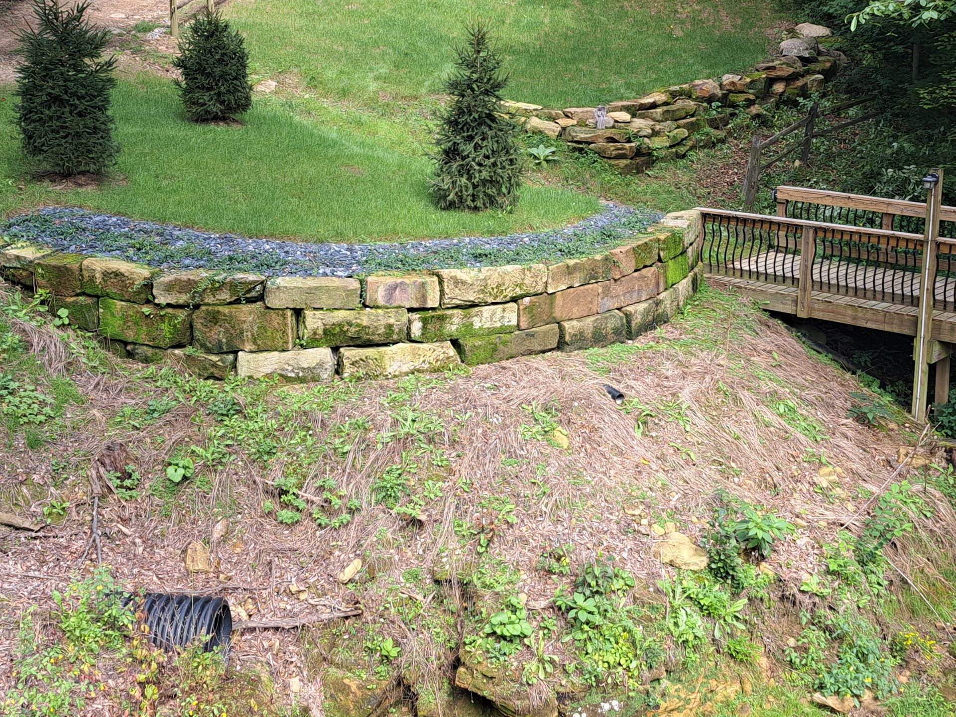 Stone retaining wall with plants, a bridge, and evergreen trees on a grassy hillside.