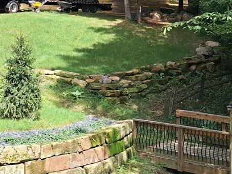 Stone retaining walls on a grassy slope with a small evergreen tree. Wooden deck with railing.