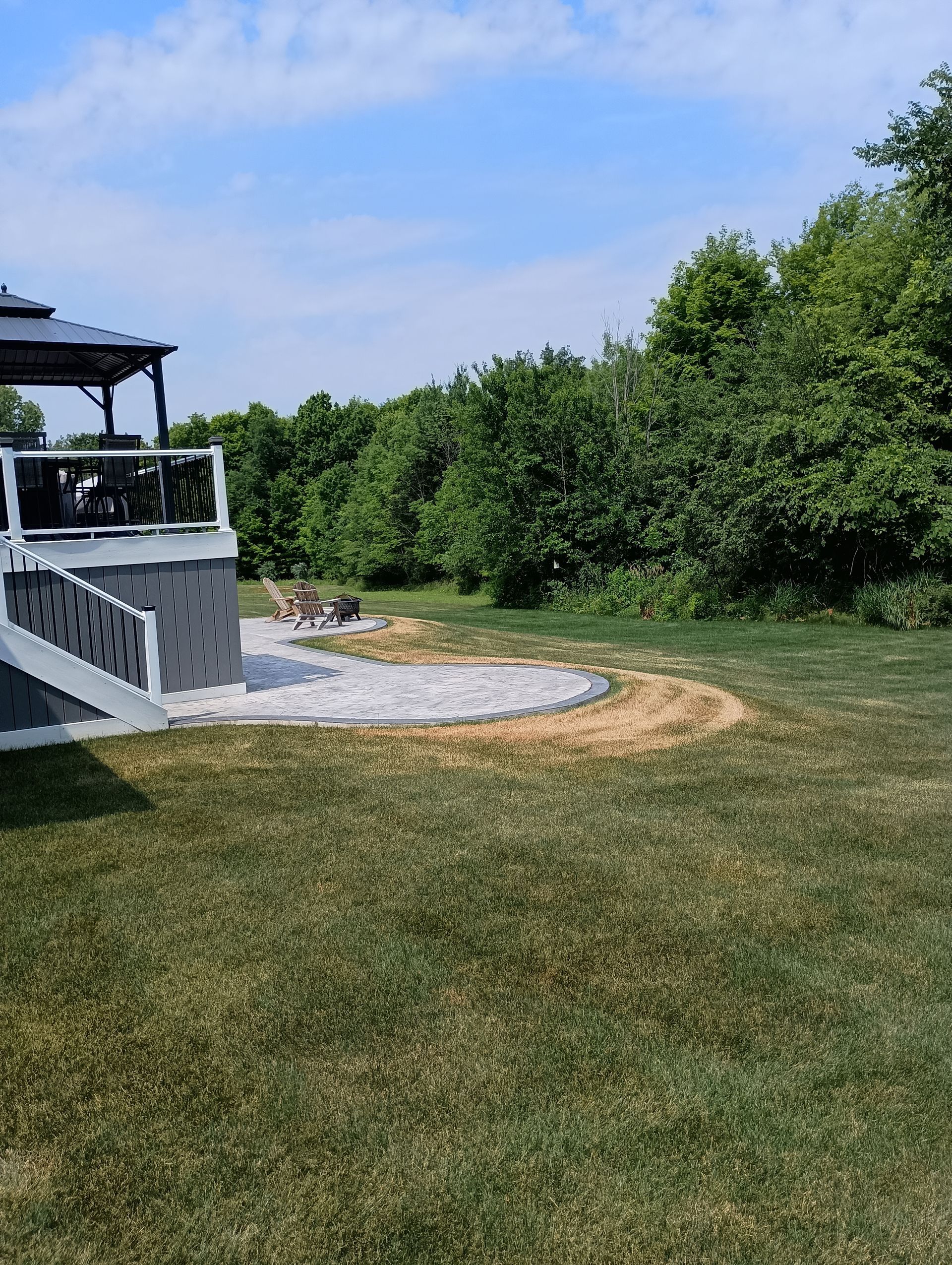 Backyard with a gray deck, stone patio, and trees under a blue sky.