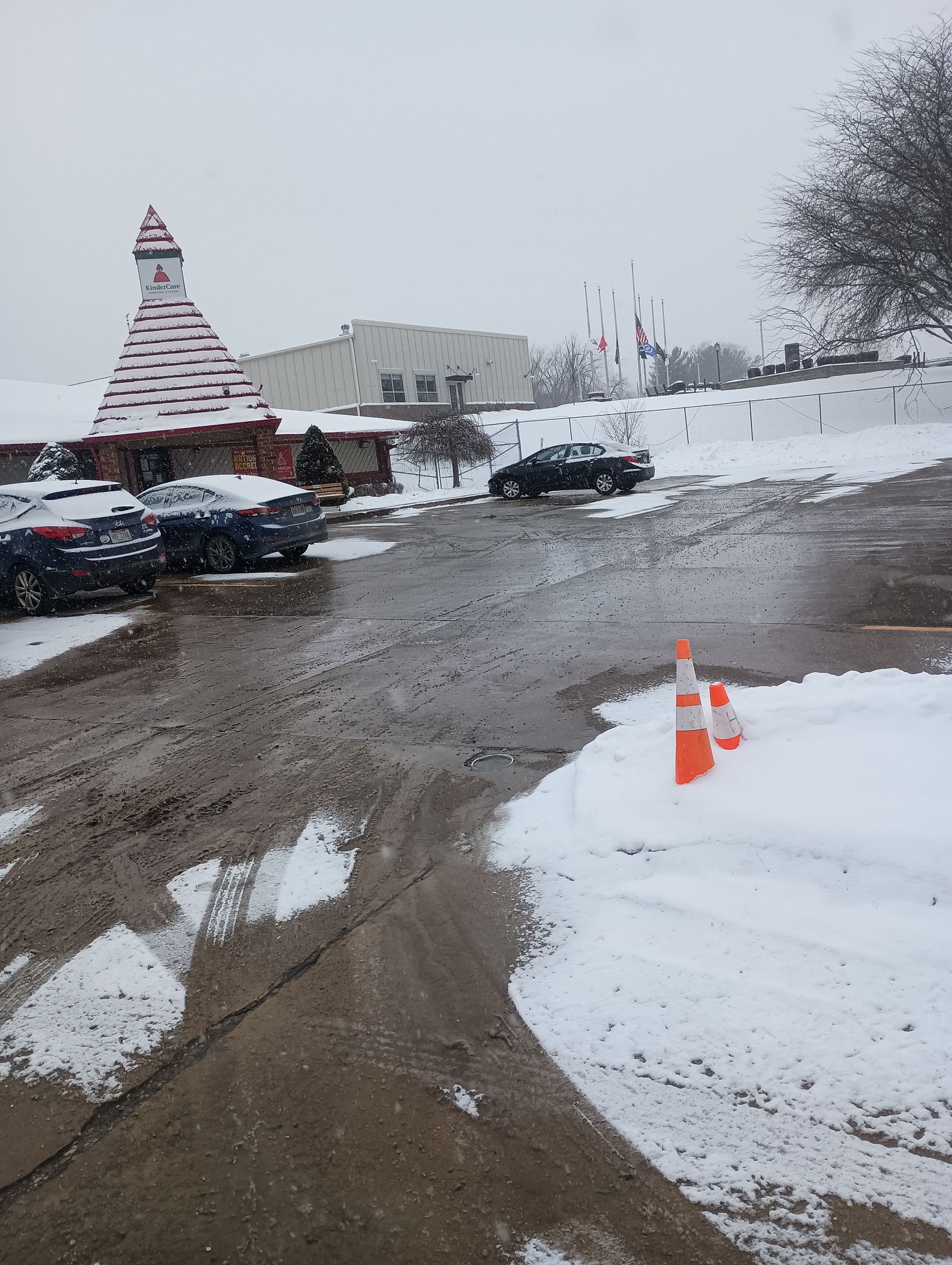 Snowy parking lot with cars and a building featuring a red and white roof; two traffic cones in the snow.