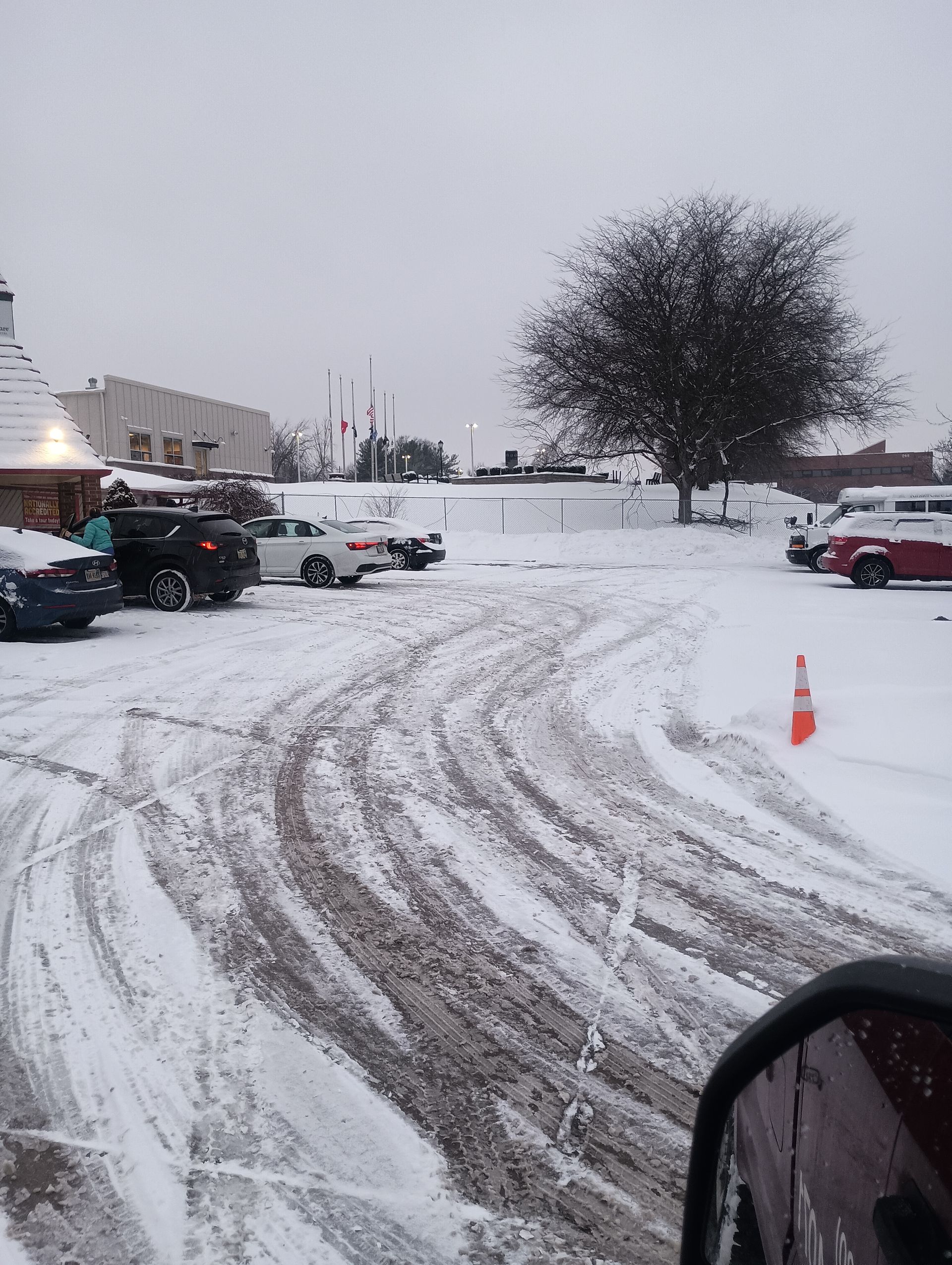 Snow-covered parking lot with tire tracks, a few cars, a tree, and a building on a cloudy day.