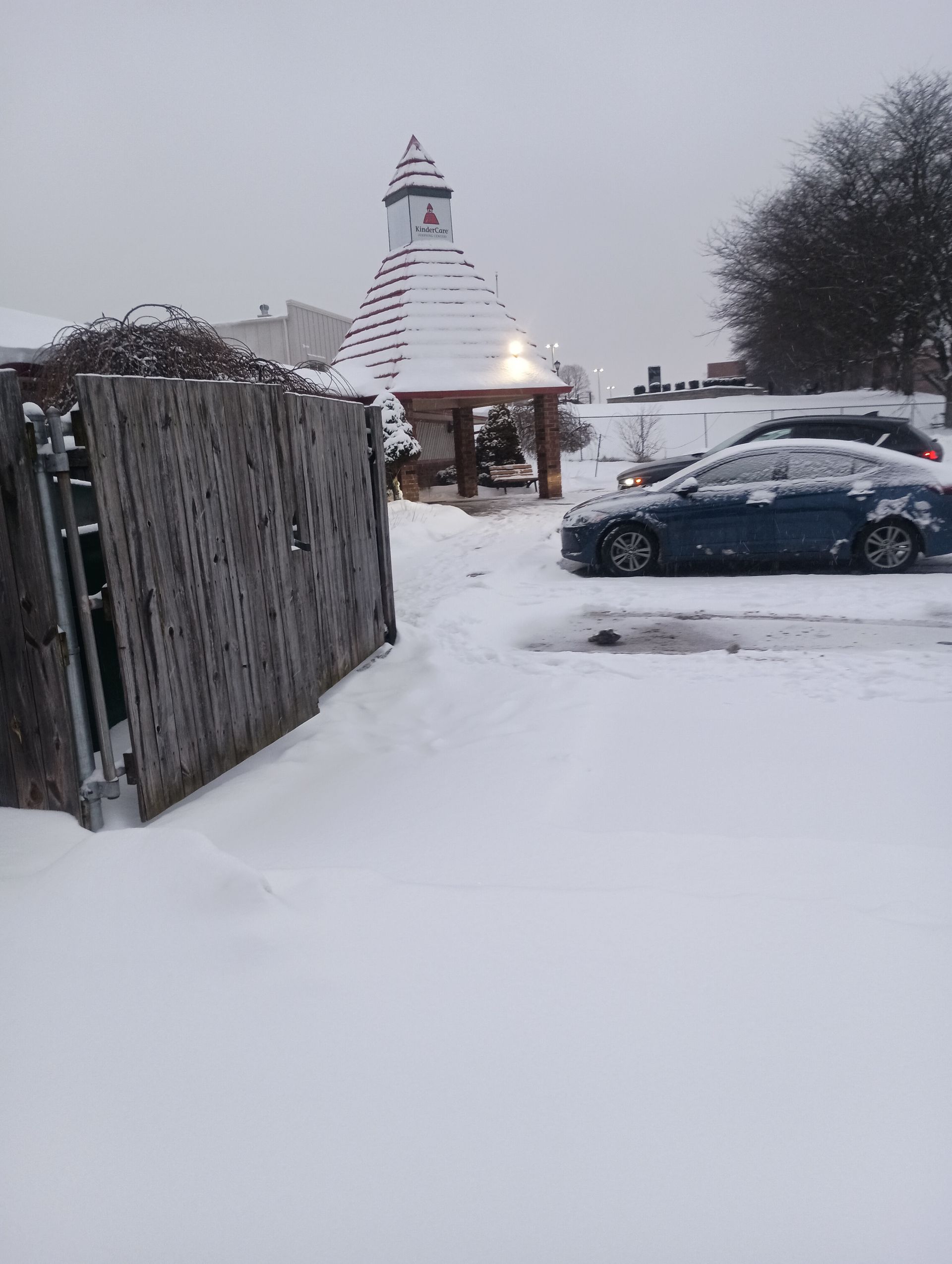Snowy scene: Wooden gazebo and cars on a snow-covered ground, near a wooden fence. Overcast sky.