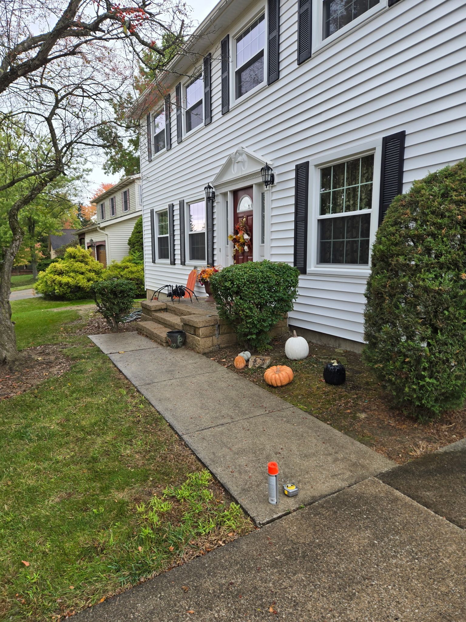 White two-story house with black shutters, concrete walkway, autumn decorations, and green lawn.