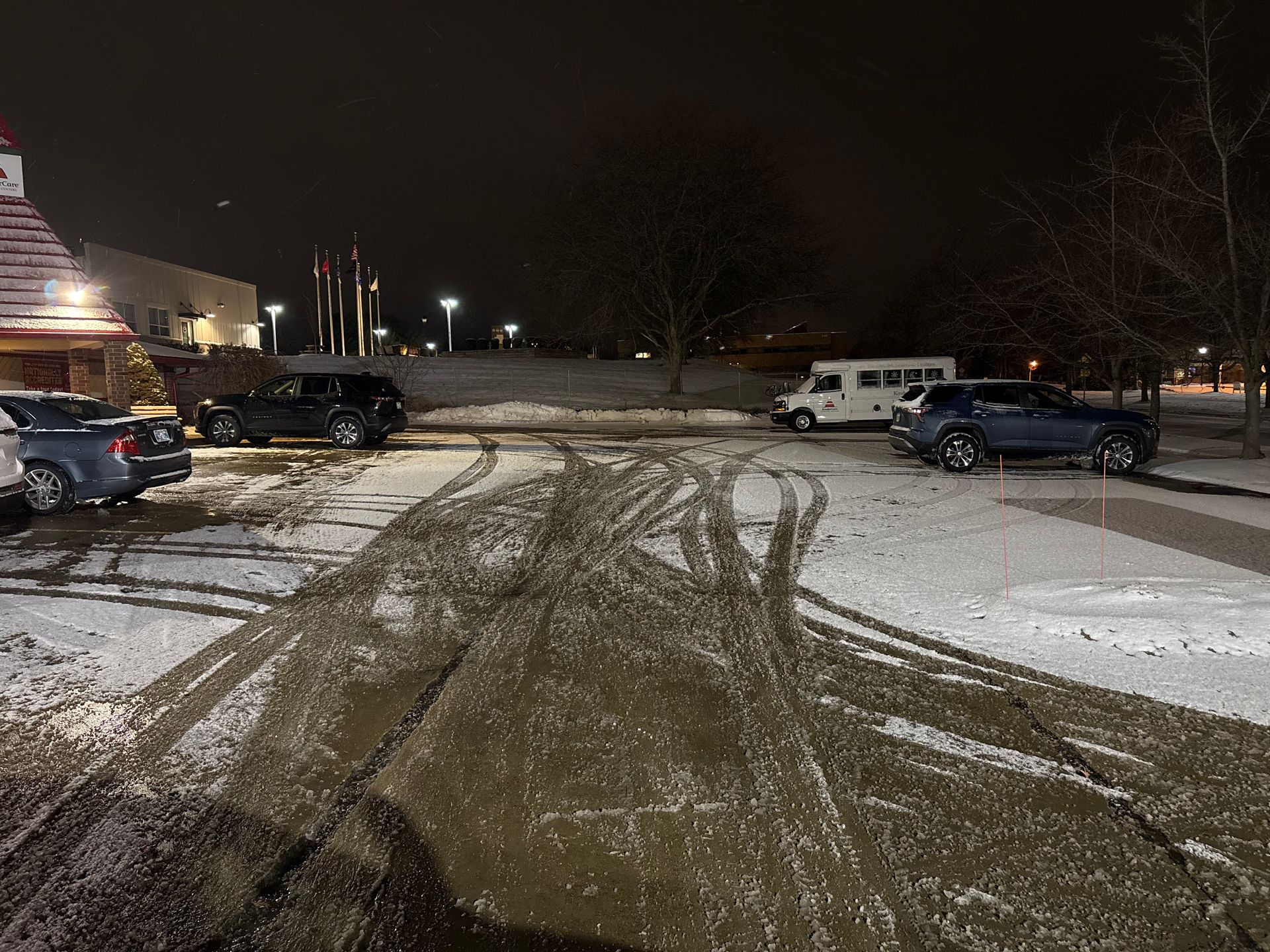 Night scene: Cars parked on a snowy lot. Muddy tire tracks in the foreground. Buildings and streetlights in the background.
