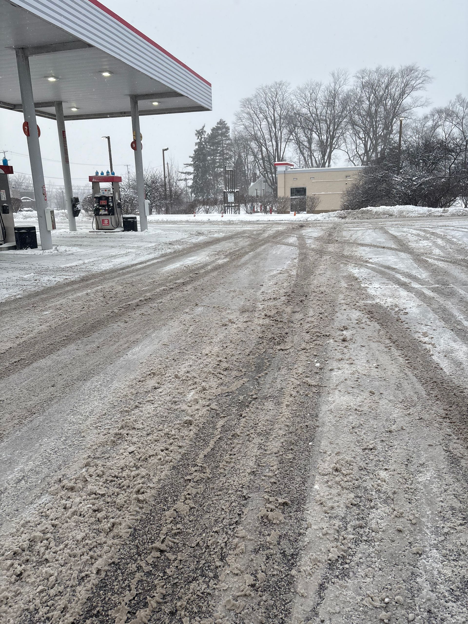 Snowy gas station scene. Tire tracks in snow-covered parking area. Canopy overhead. Bare trees in background.