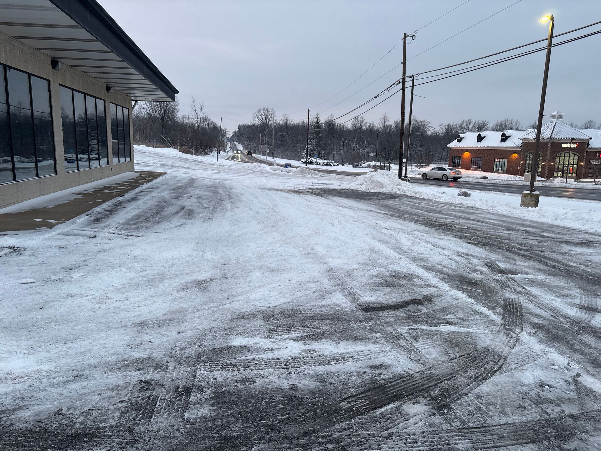 Snowy parking lot with building on the left and a building with lights on the right. A person walks in the distance.