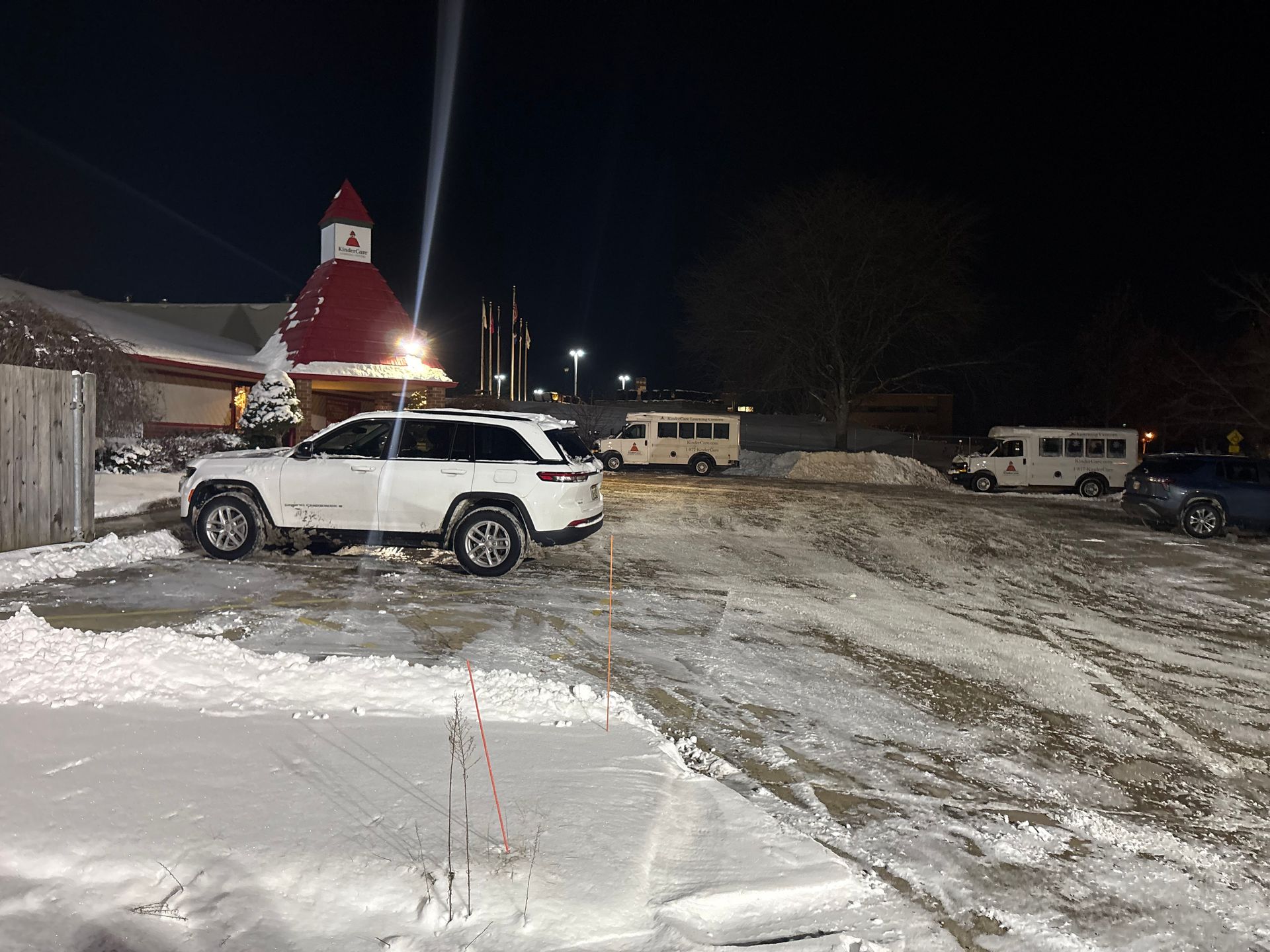 Snowy night scene with a white SUV, a building with a red roof, and two small white buses parked in a snow-covered lot.