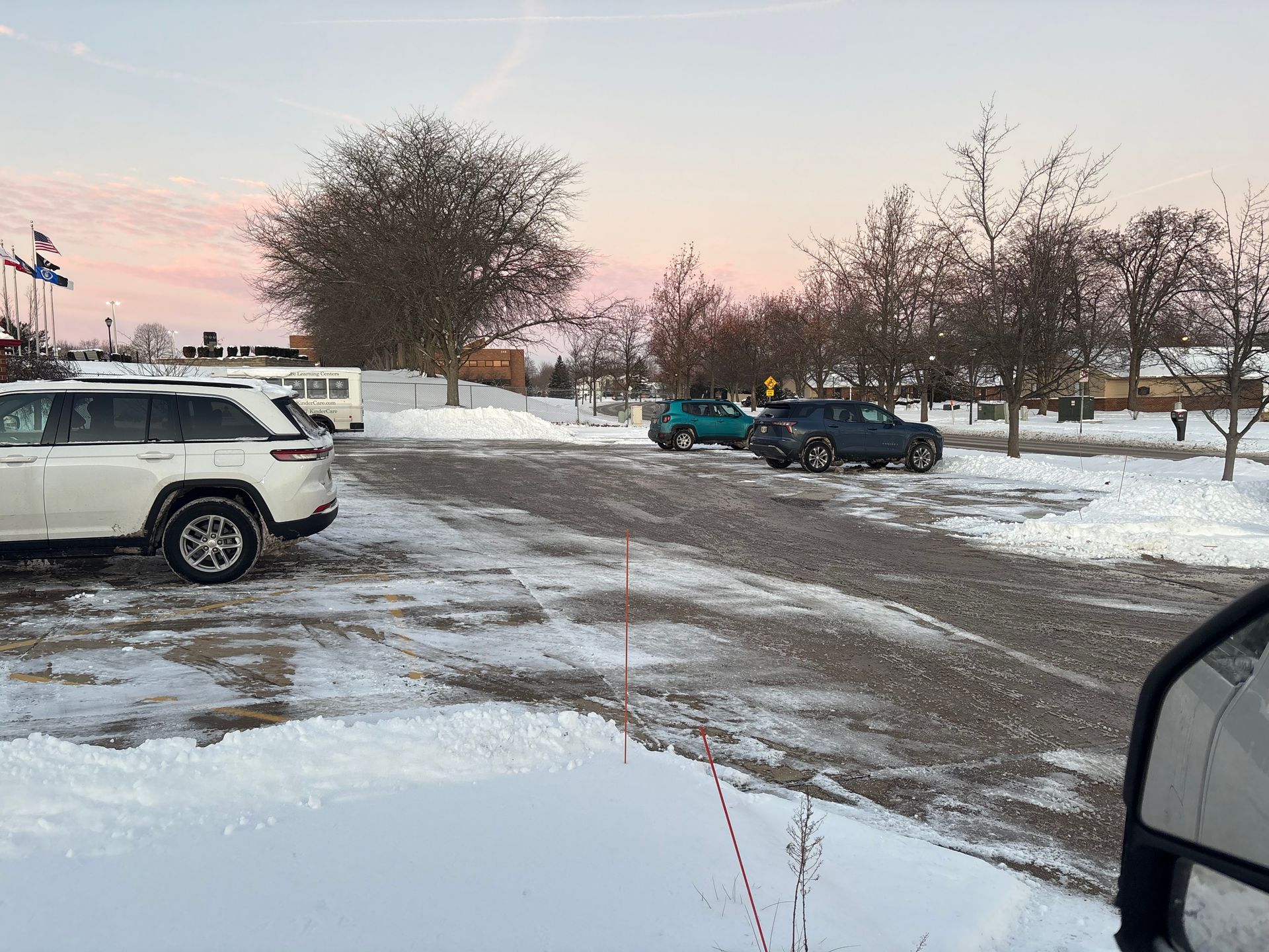Snowy parking lot with cars and bare trees. Sky is pink and gray.
