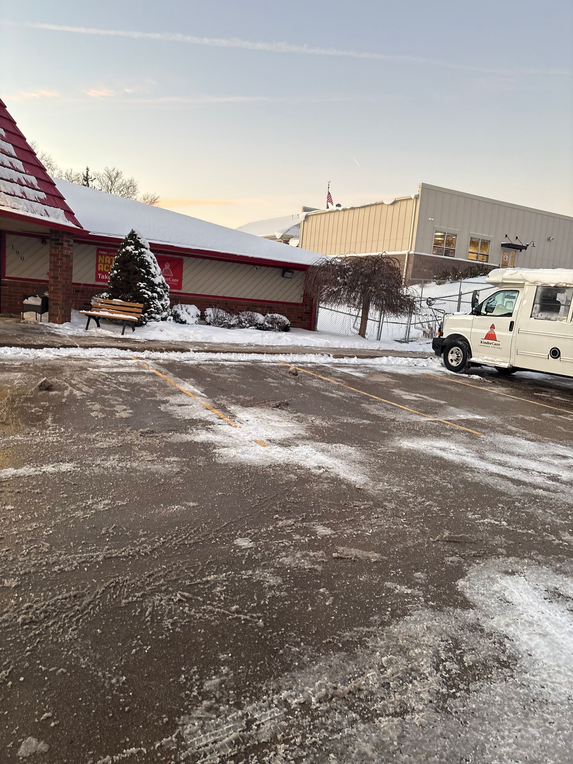 Snow-covered parking lot with a low building, evergreen tree, and utility truck on a winter day.