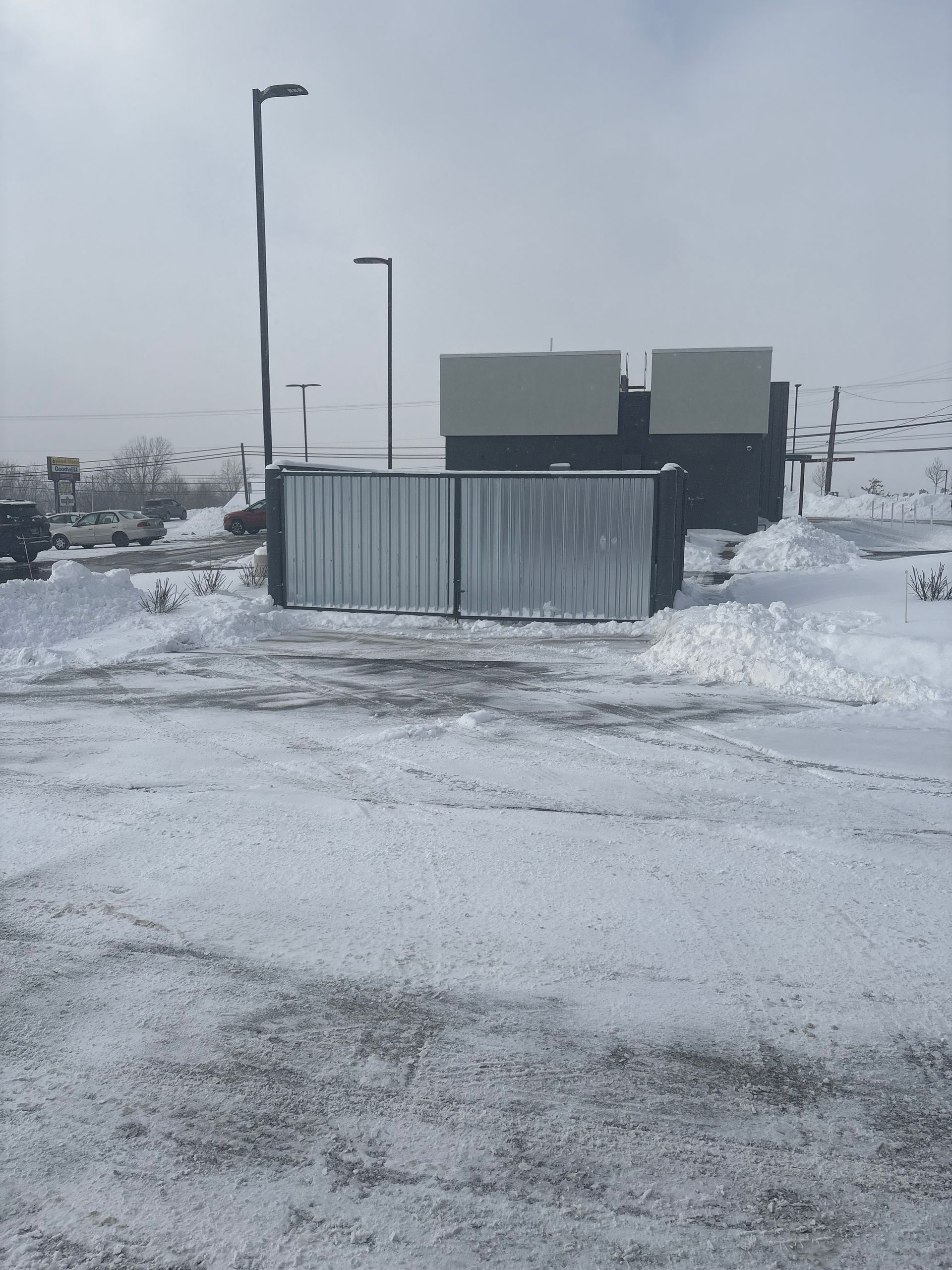 Snowy scene: Metal gate and building with snow-covered ground. Streetlights and overcast sky.