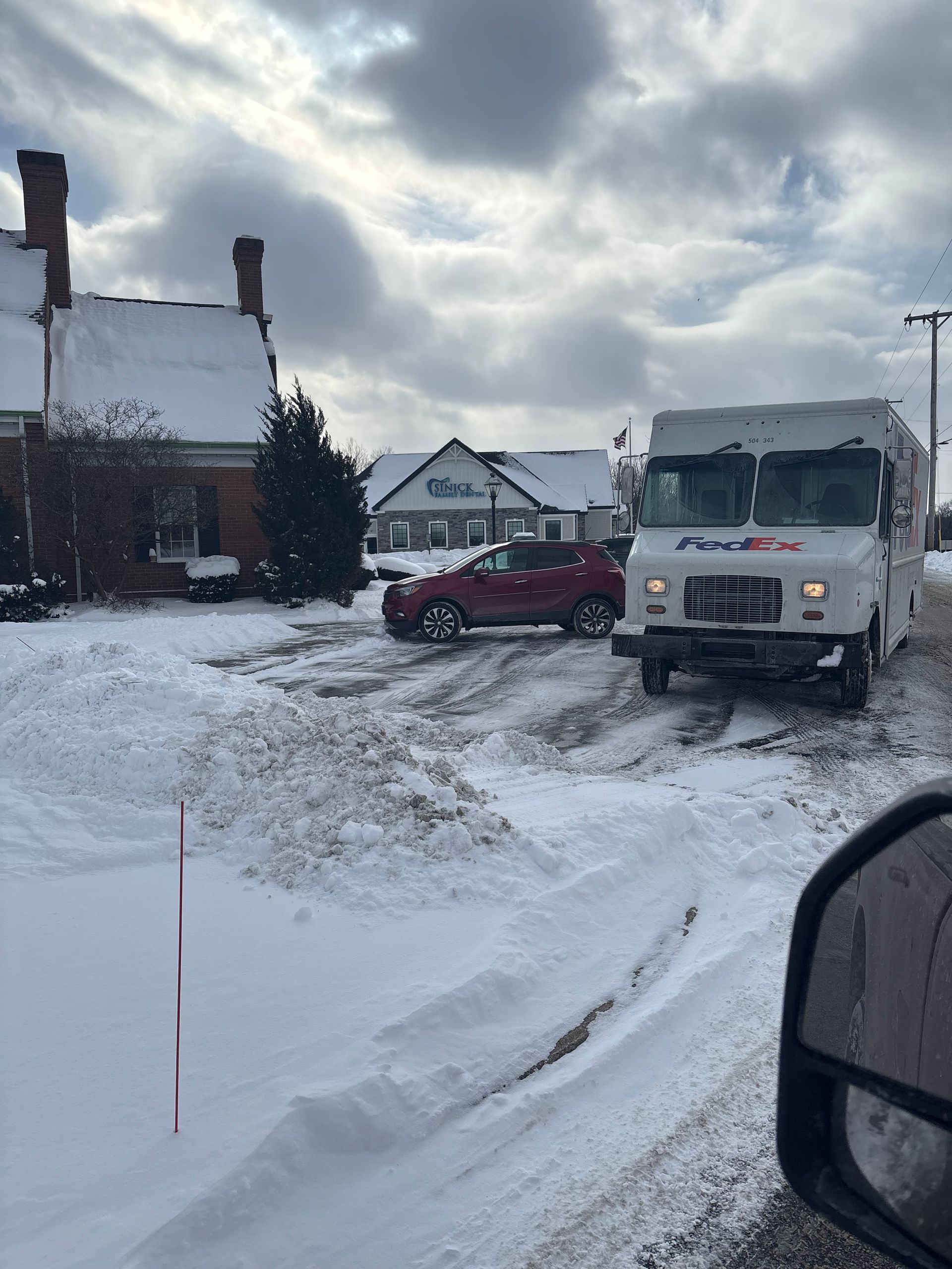 Snowy scene: Red SUV and white truck stuck in snow, near buildings. Cloudy sky.