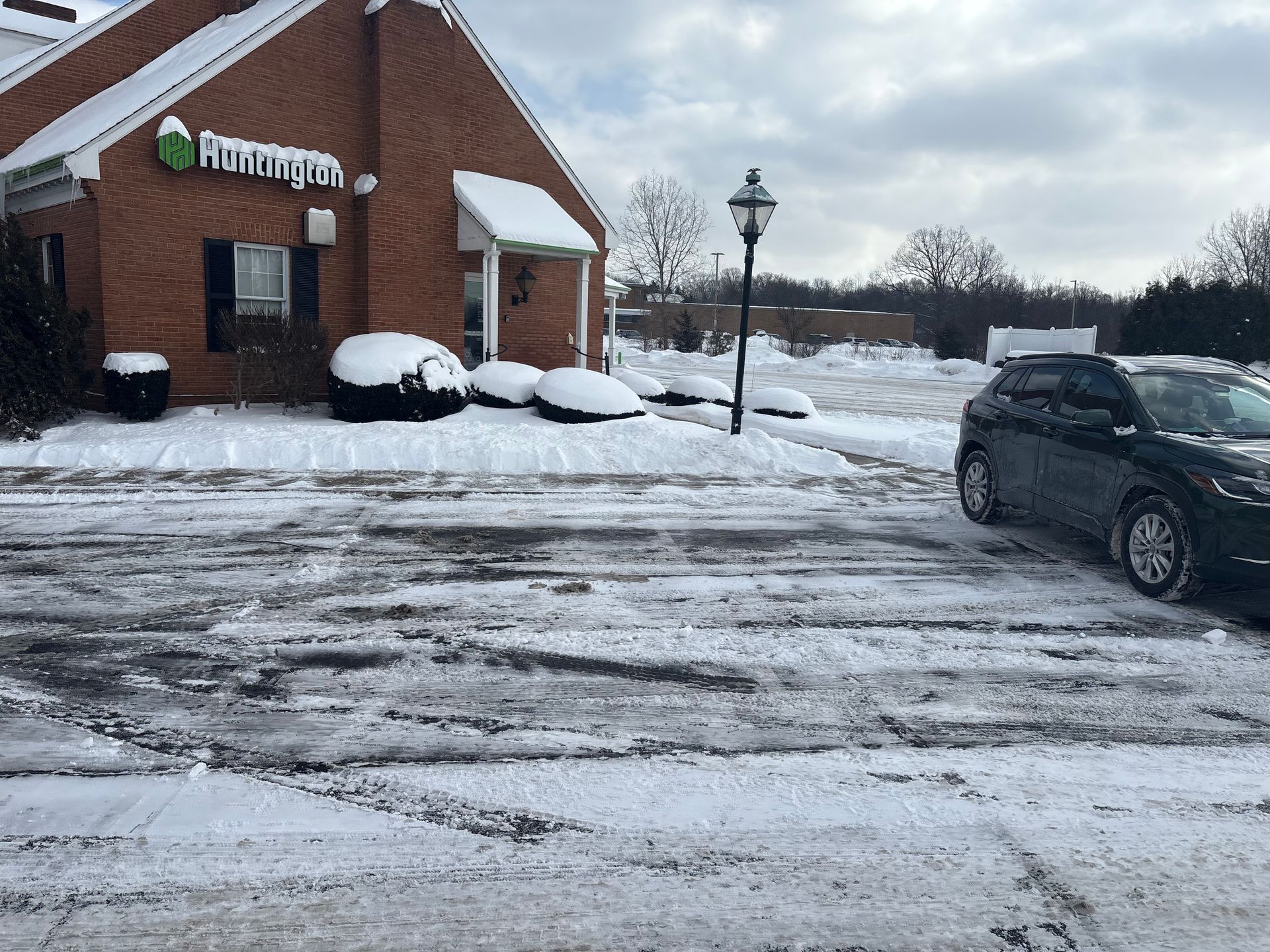 Snowy parking lot in front of a brick building with a sign, a parked car on the right.