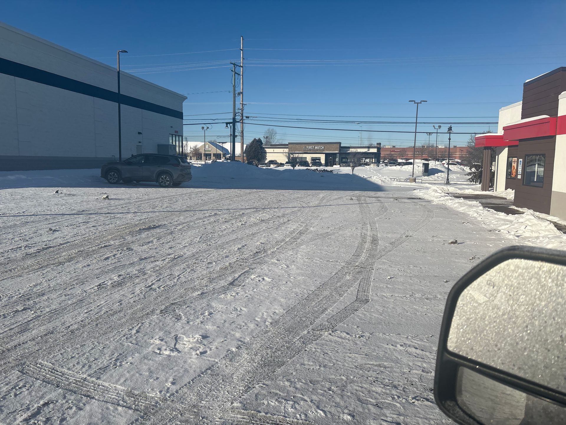 Snowy parking lot with car tracks, industrial building left, fast food restaurant right, sunny day.