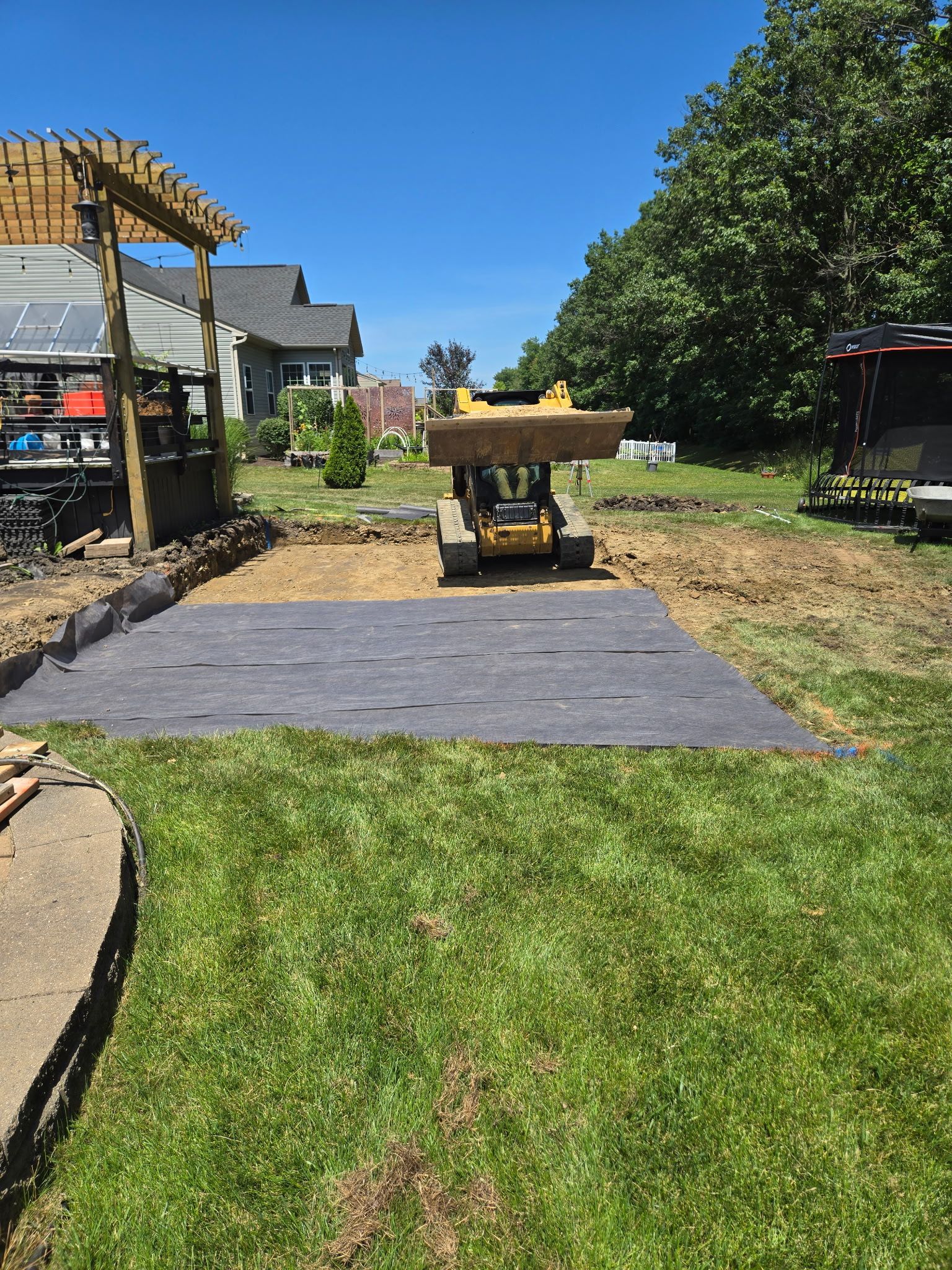 A small construction vehicle on gravel with landscaping fabric, in a backyard. A pergola and house are visible.