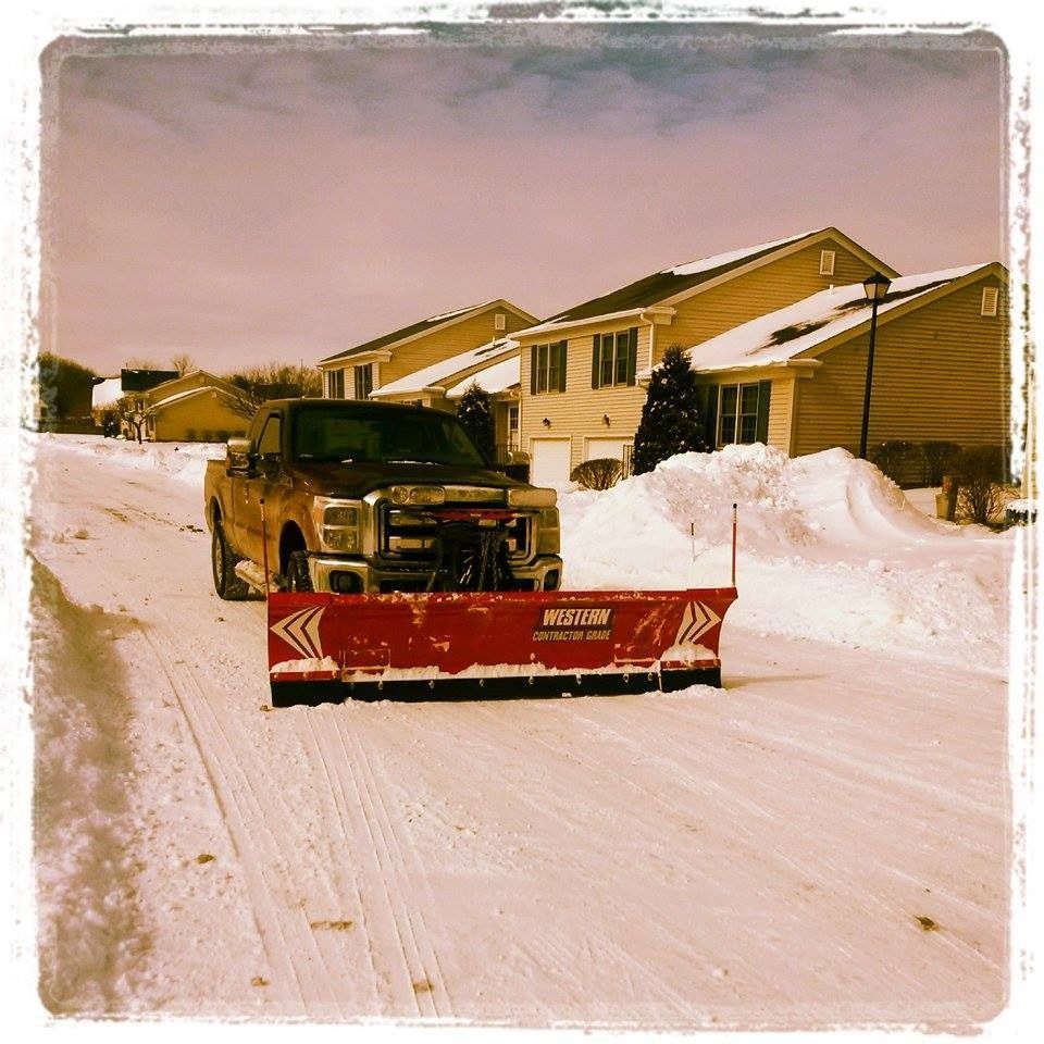 A truck with a red snowplow clears a snow-covered street in a residential area.