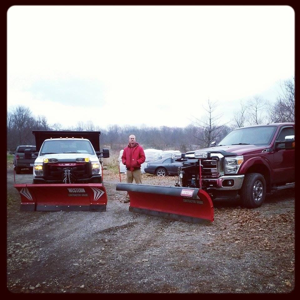 Man standing between two red pickup trucks with snowplows attached. Overcast day.