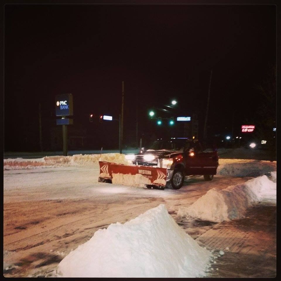Snowplow clearing a snowy parking lot at night with a PNC Bank sign in the background.