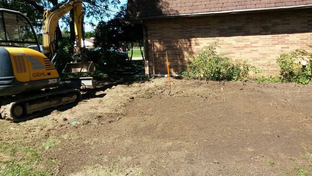 A small excavator digs near a brick building. Earth and grass visible.