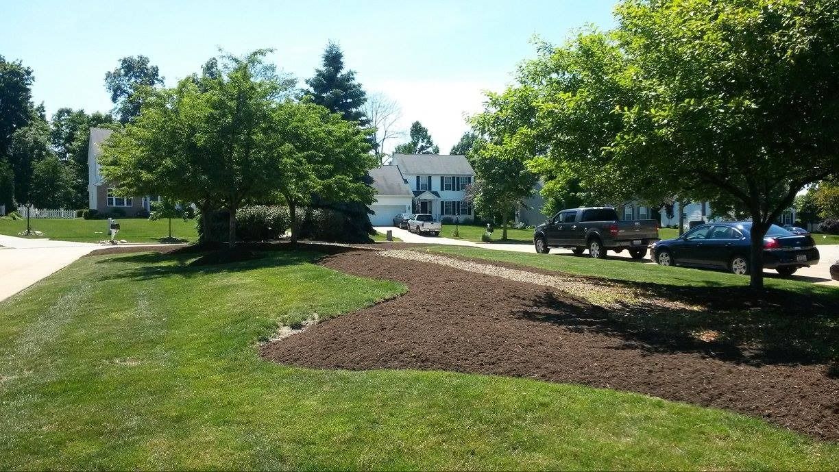 Green lawn with mulch bed and trees in front of houses; cars parked on street.