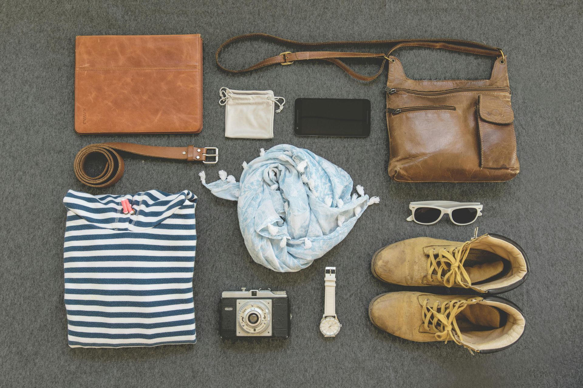 Plano de accesorios de viaje: bolso de cuero, botas, cámara, bufanda, reloj, camisa, billetera y gafas de sol en gris.