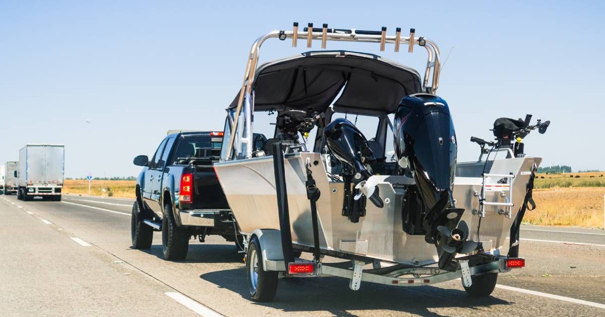 A black truck is towing a large boat. The truck is driving on a highway with two semi-trucks in the 