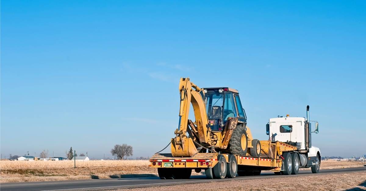 A semi-truck is hauling a large backhoe loader on a sunny day. The truck is driving down a remote street.