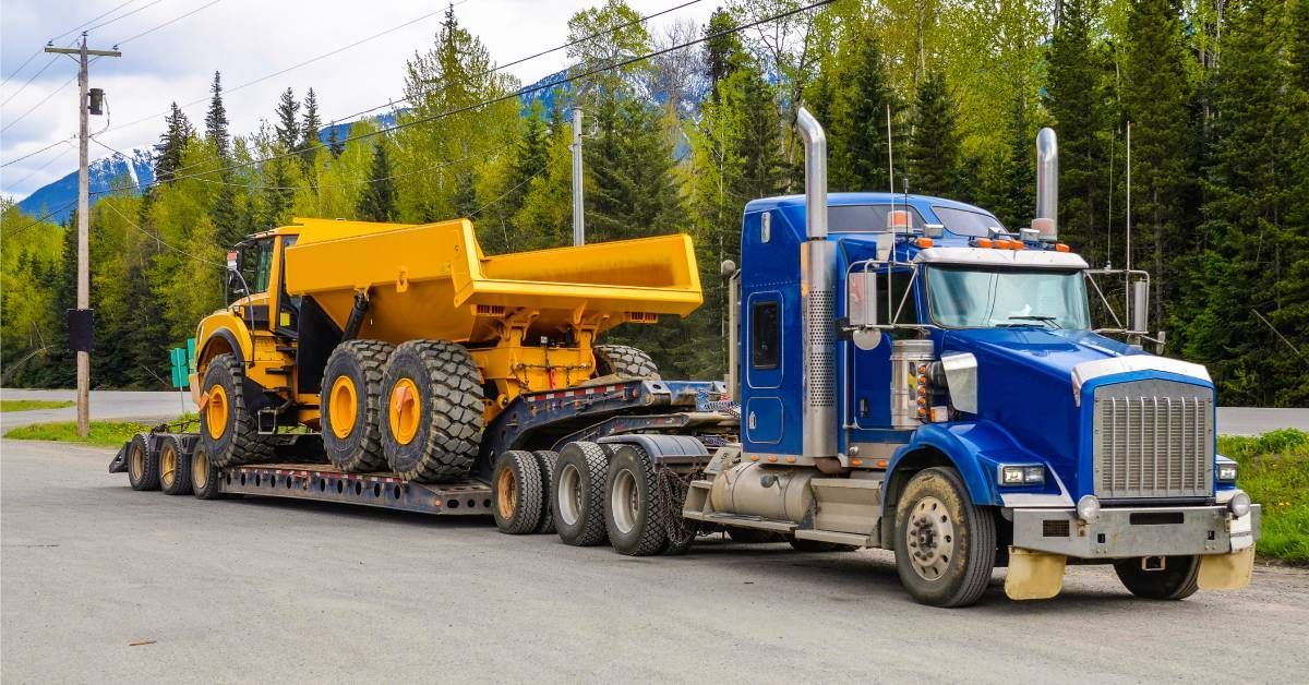 A large truck with a low platform trailer is hauling a tipper truck. The truck is driving down a tree-lined street.