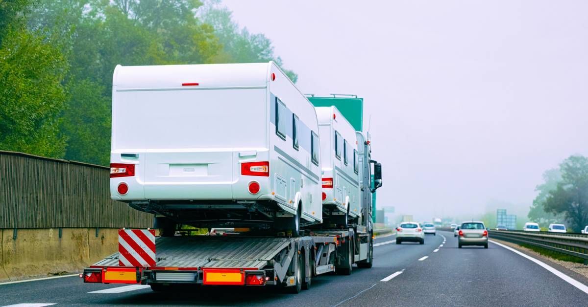 A semi-truck transporting two white motor homes along a highway. There are cars driving in front of the truck.