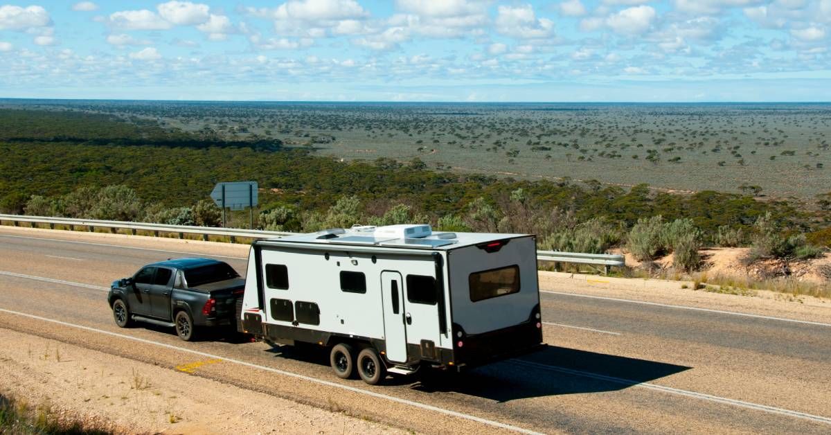 A pickup truck hauling a large white RV on a highway. The road is surrounded by shrubs and open land