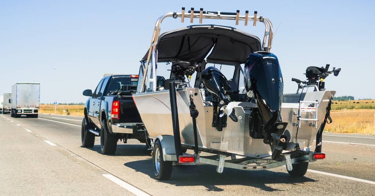 A pickup truck towing a silver boat on a highway. There are two semi-trucks in front of the pickup.